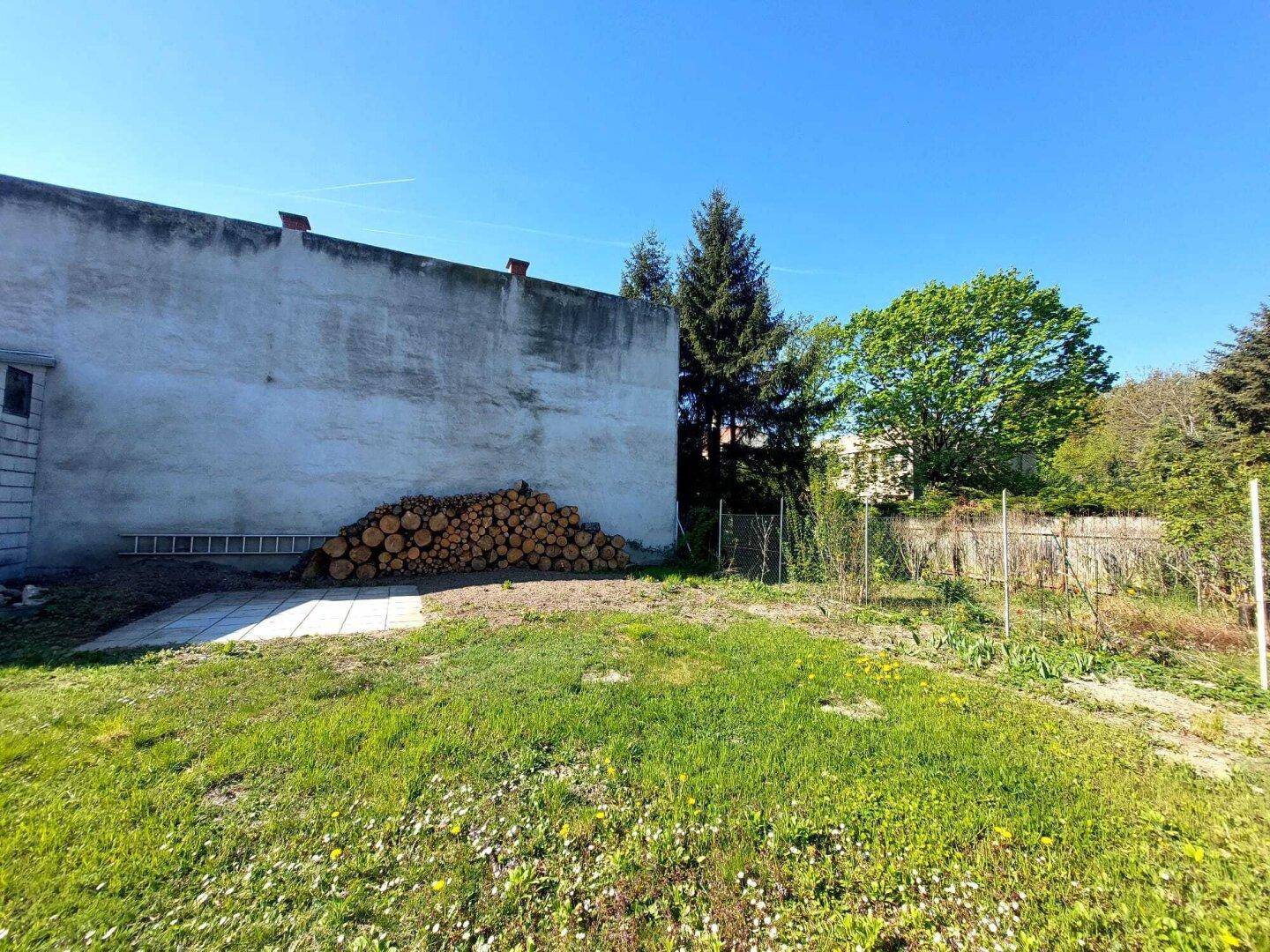 Gartenbereich mit Holzstapel, kleiner gepflasterter Fläche und grüner Wiese neben einer Gebäudewand.