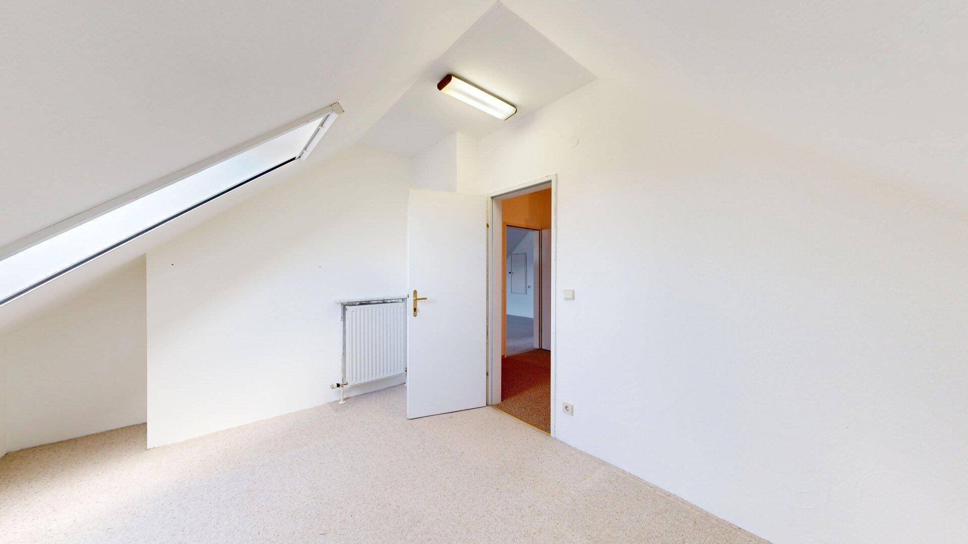 Helles Dachzimmer mit Teppichboden, Dachschräge und Heizkörper unter dem Fenster.
