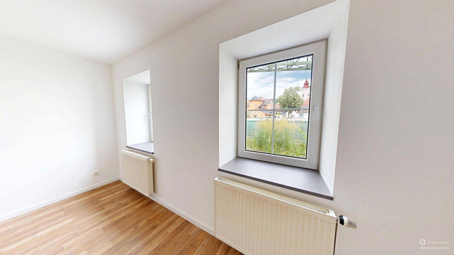 Helles Zimmer mit Laminatboden und zwei Fenstern, die einen Blick ins Grüne bieten.