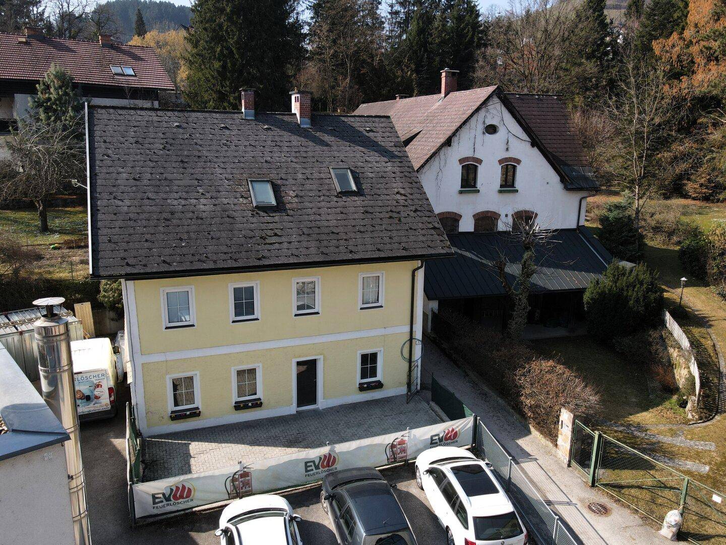 Ansicht eines gelben Wohnhauses mit dunklem Dach und Parkplätzen im Vordergrund.