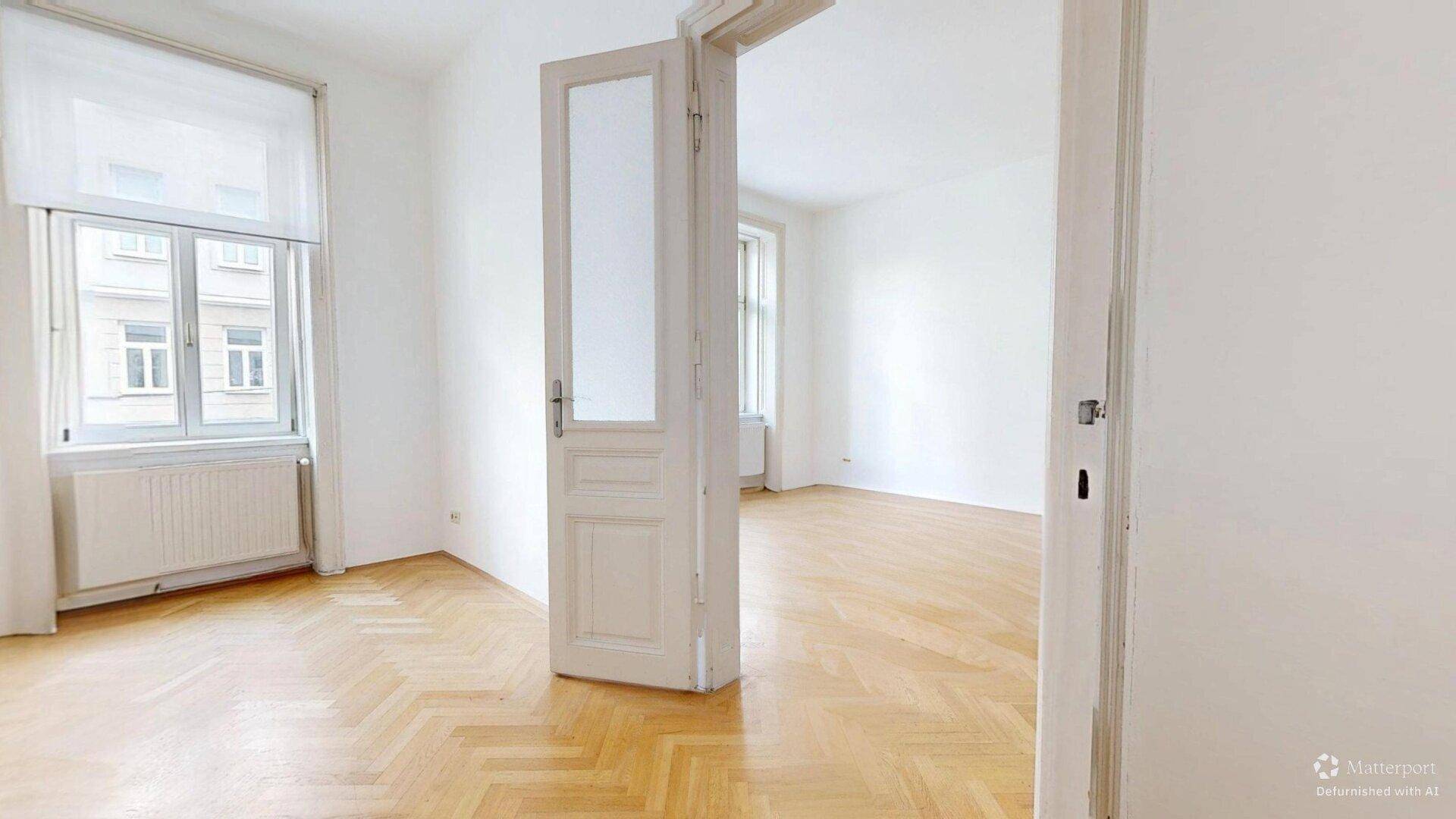 Helles Zimmer mit Fischgrätparkett und einer Flügeltür, die zu einem weiteren Raum führt.