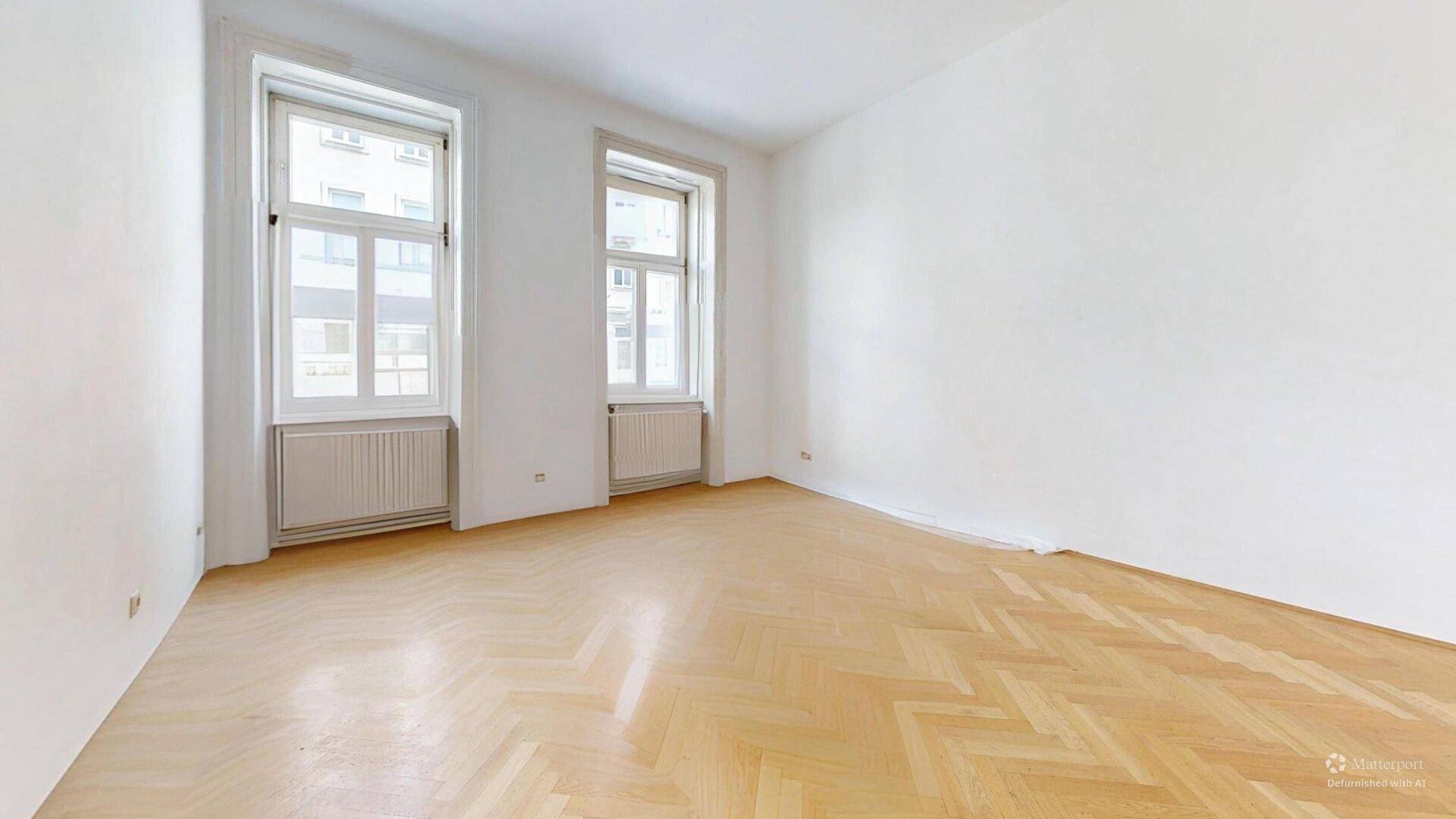 Helles, leeres Zimmer mit Fischgrätparkett und zwei hohen Fenstern, ideal für Wohnzwecke.