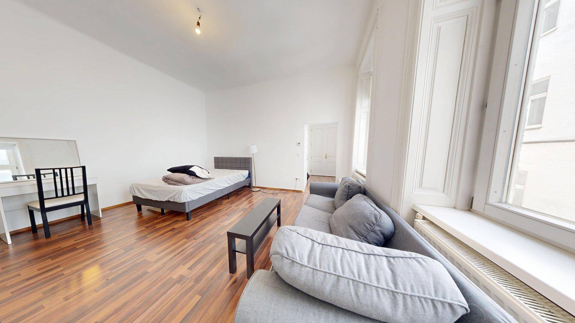 Möbliertes Schlafzimmer mit Bett, Sofa, Schminktisch und großen Fenstern, die viel Licht hereinlassen.