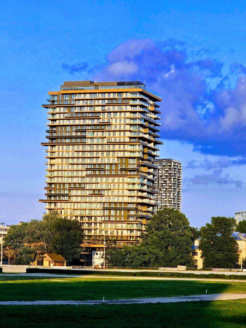 Modernes Hochhaus mit vielen Balkonen und Glasfassade, umgeben von Grünflächen unter blauem Himmel.