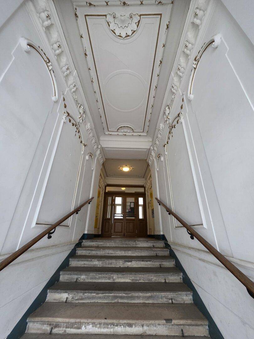 Eindrucksvolles Treppenhaus mit hohen Decken, kunstvollen Stuckverzierungen und Steinstufen in einem Altbau.