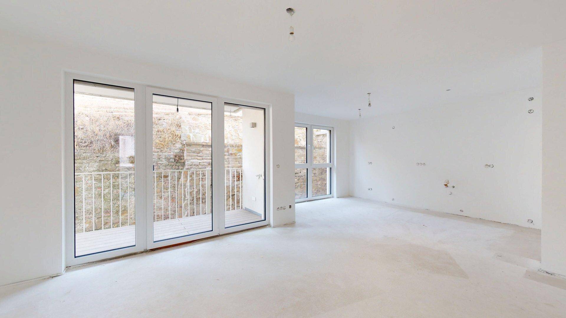 Heller, unmöblierter Innenraum im Rohbau mit großen Fenstern und Zugang zu einem kleinen Balkon.