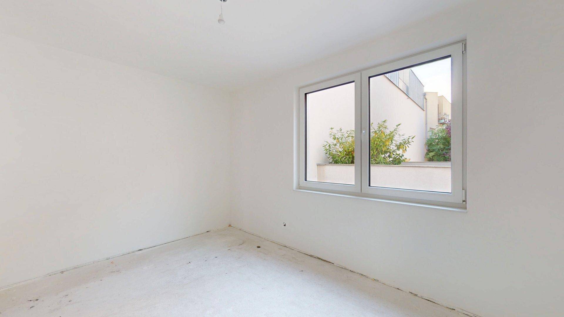 Helles Schlafzimmer im Rohbau mit einem großen Fenster und Blick auf eine begrünte Außenanlage.