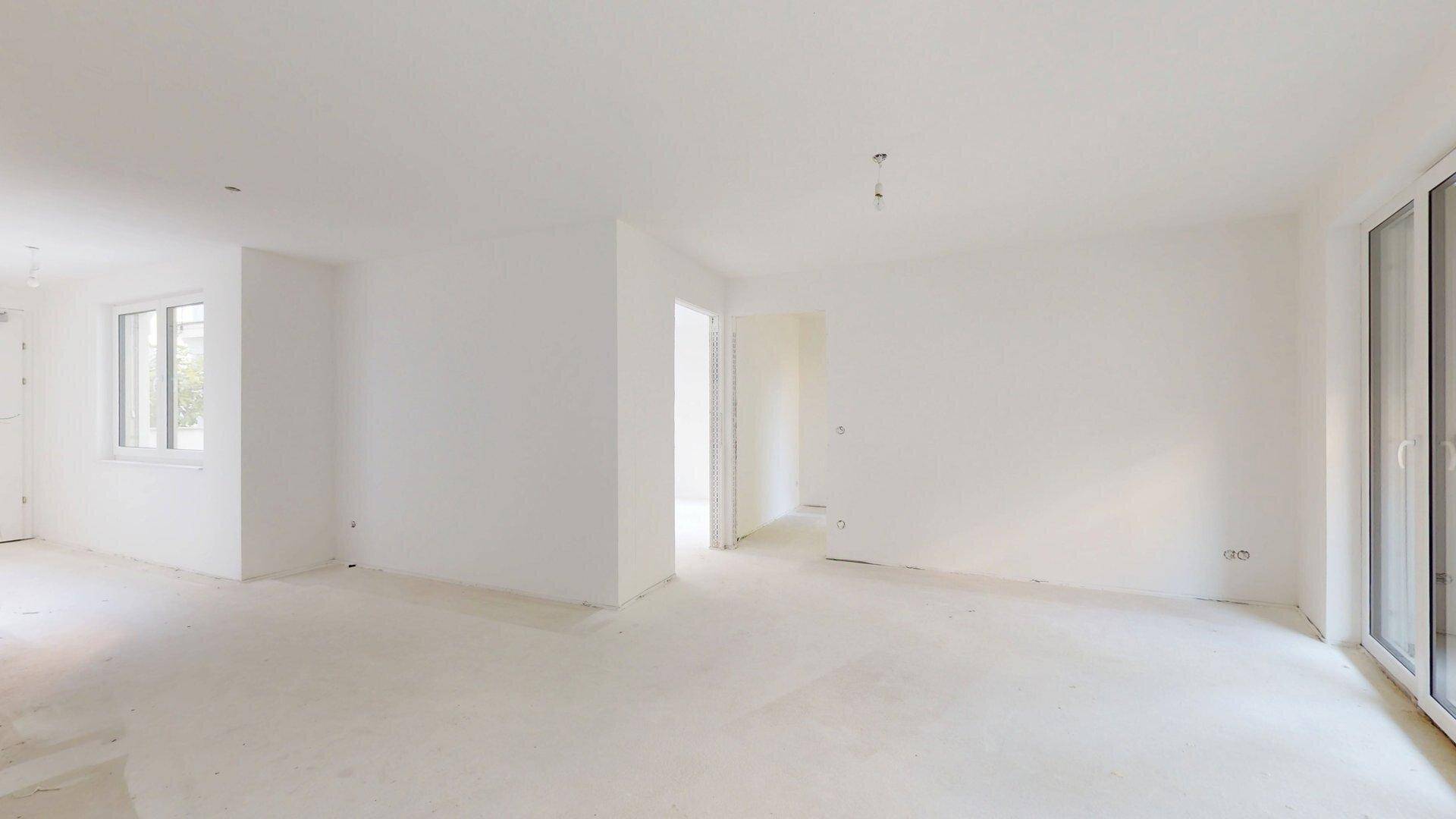 Heller, unmöblierter Wohnbereich im Rohbau mit großen Fenstern und viel natürlichem Lichteinfall.