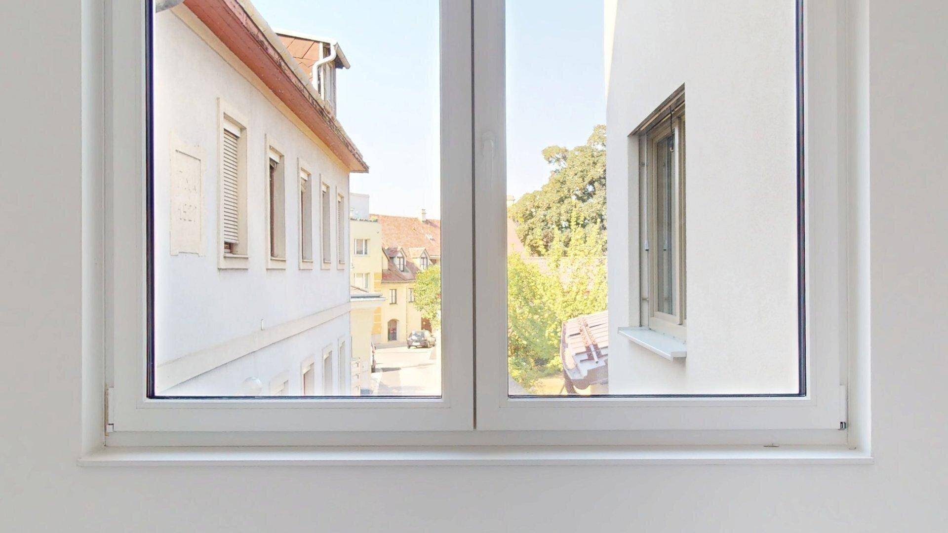 Nahaufnahme eines Fensters mit Blick auf die Straße, umliegende Gebäude und grüne Bäume.