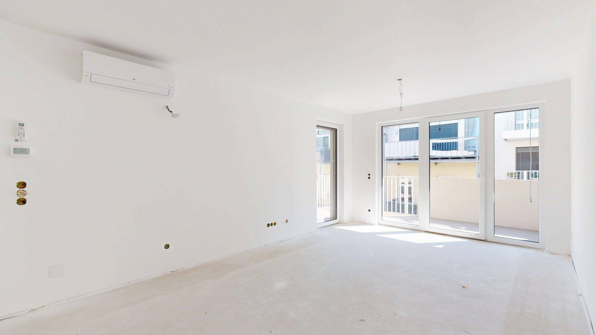 Unfertiger Wohnbereich mit weißen Wänden, vorbereiteten Anschlüssen und Zugang zum Balkon.