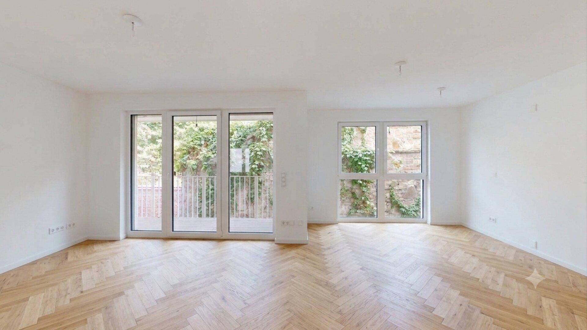 Heller Wohnbereich mit Fischgrätparkett, großen Fenstern und Zugang zu einem Balkon mit Blick ins Grüne.