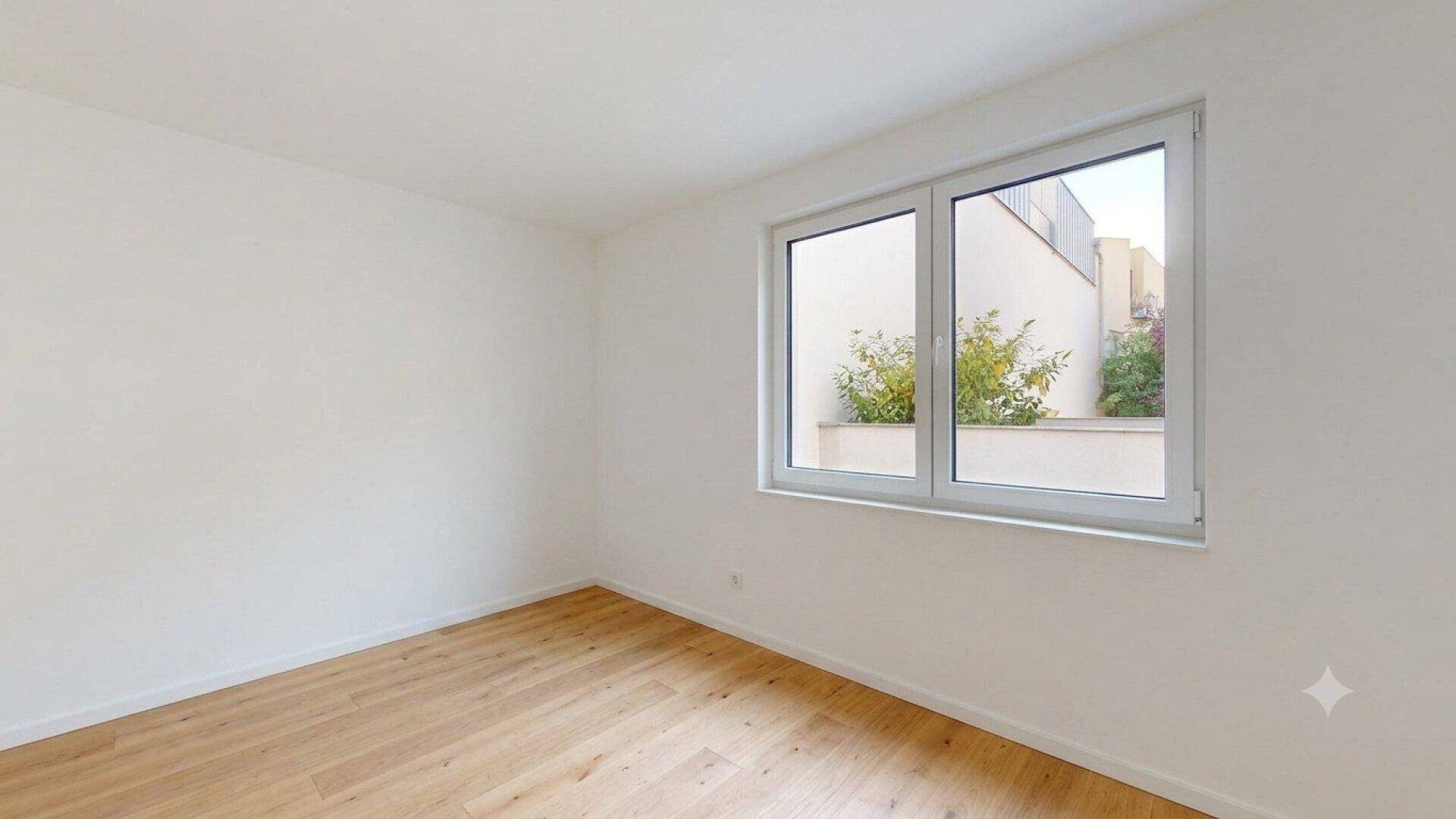 Helles, leeres Zimmer mit großem Fenster und Blick auf die umliegende Bebauung und Grünflächen.
