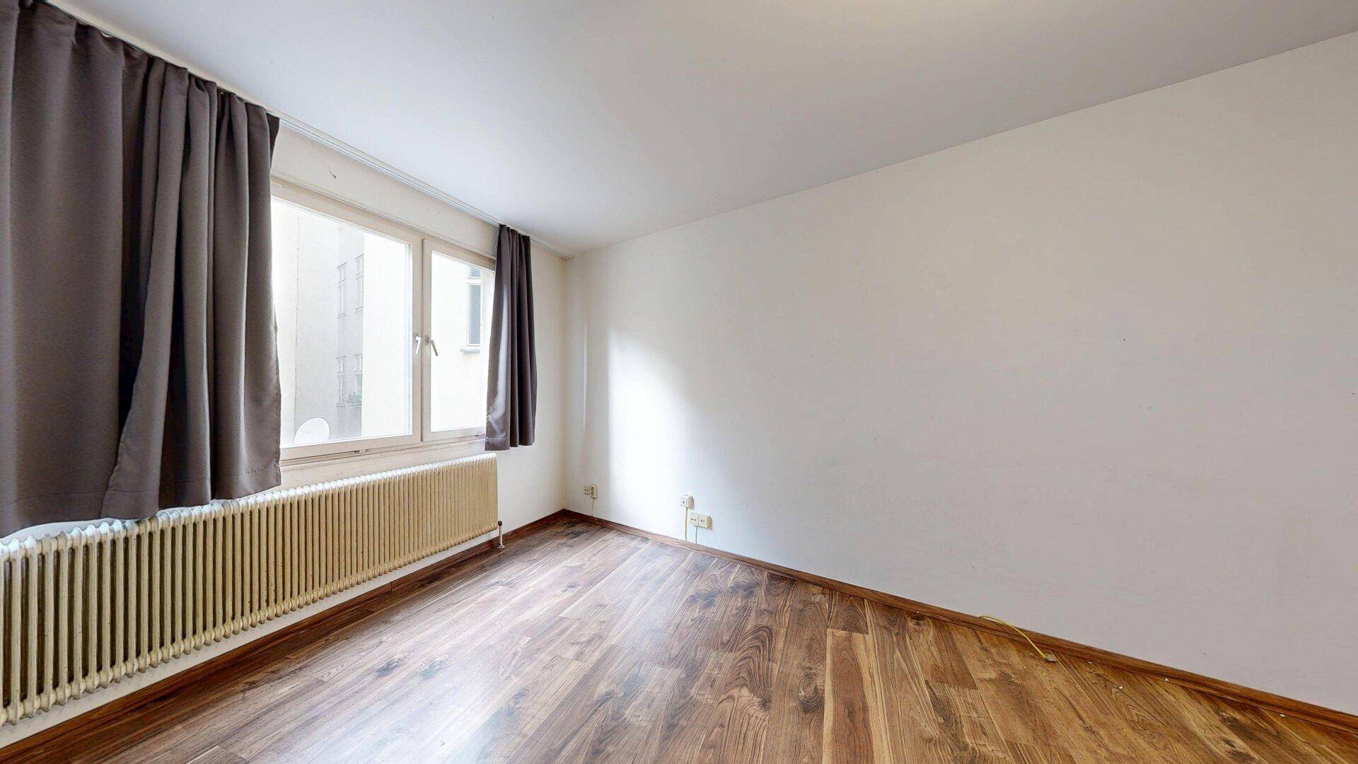 Geräumiges Zimmer mit Holzboden, weißen Wänden und einem großen Fenster mit dunklen Vorhängen.