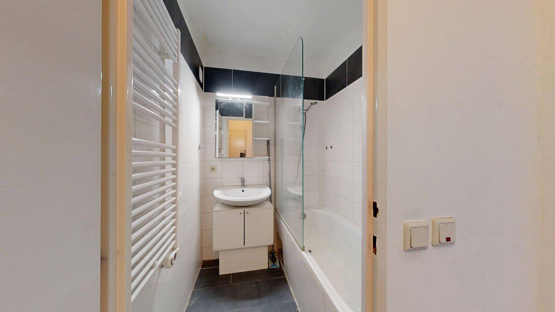 Badezimmer mit Waschbecken, Spiegel, Badewanne und Handtuchheizkörper, durch eine Tür sichtbar.