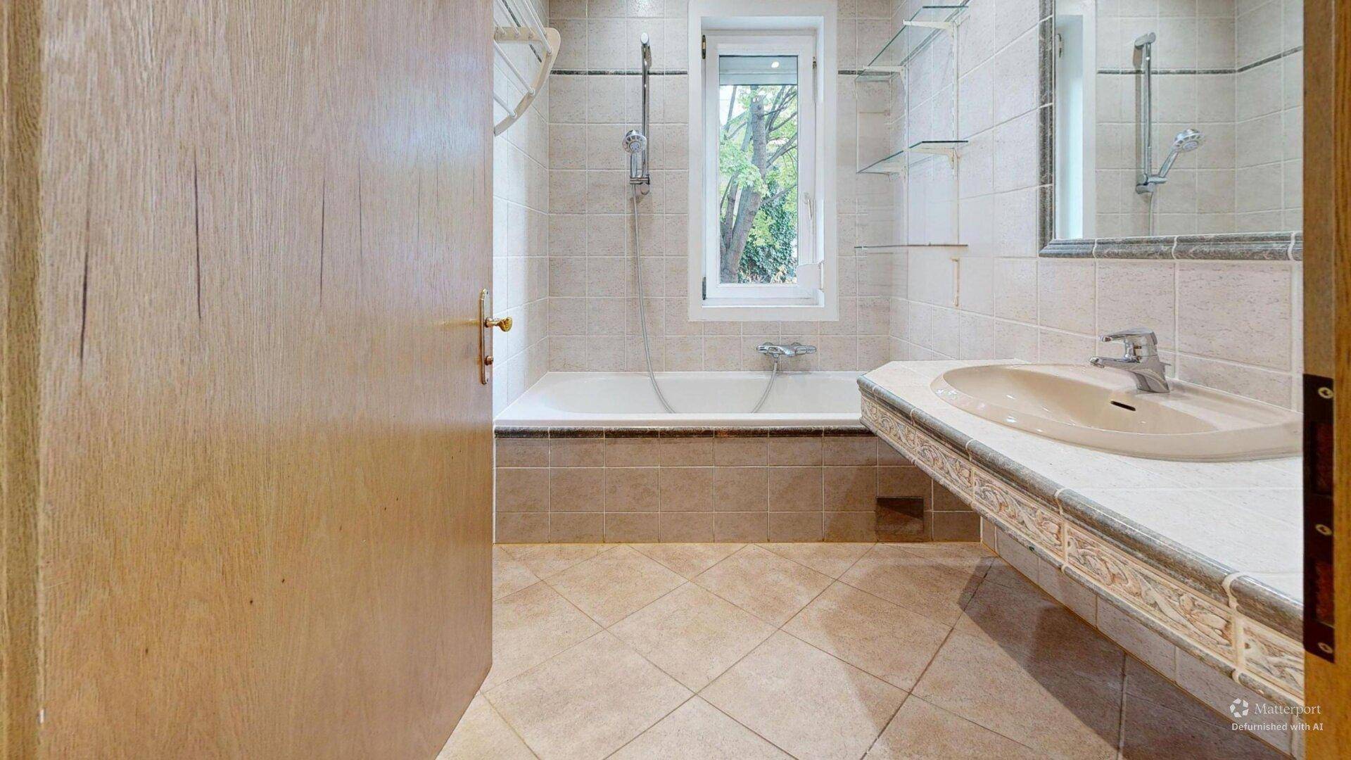Helles Badezimmer mit beigefarbenen Fliesen, Badewanne und Waschtisch, durch eine Holztür zugänglich.