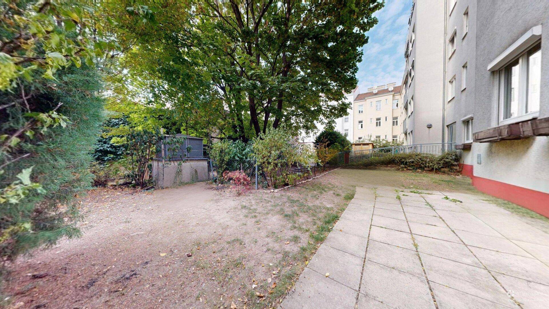 Ungepflegter Gartenbereich mit Bäumen und einem gepflasterten Weg neben einem Wohngebäude.
