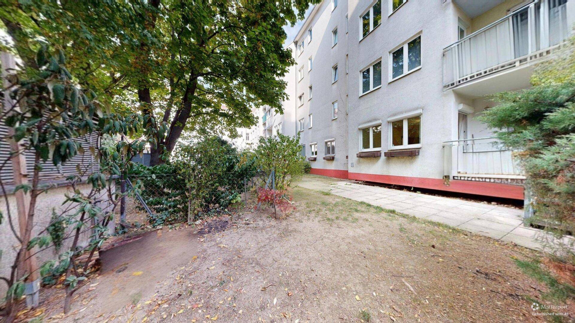 Schmaler, naturbelassener Gartenstreifen entlang der Gebäudefassade mit Bäumen und Sträuchern.