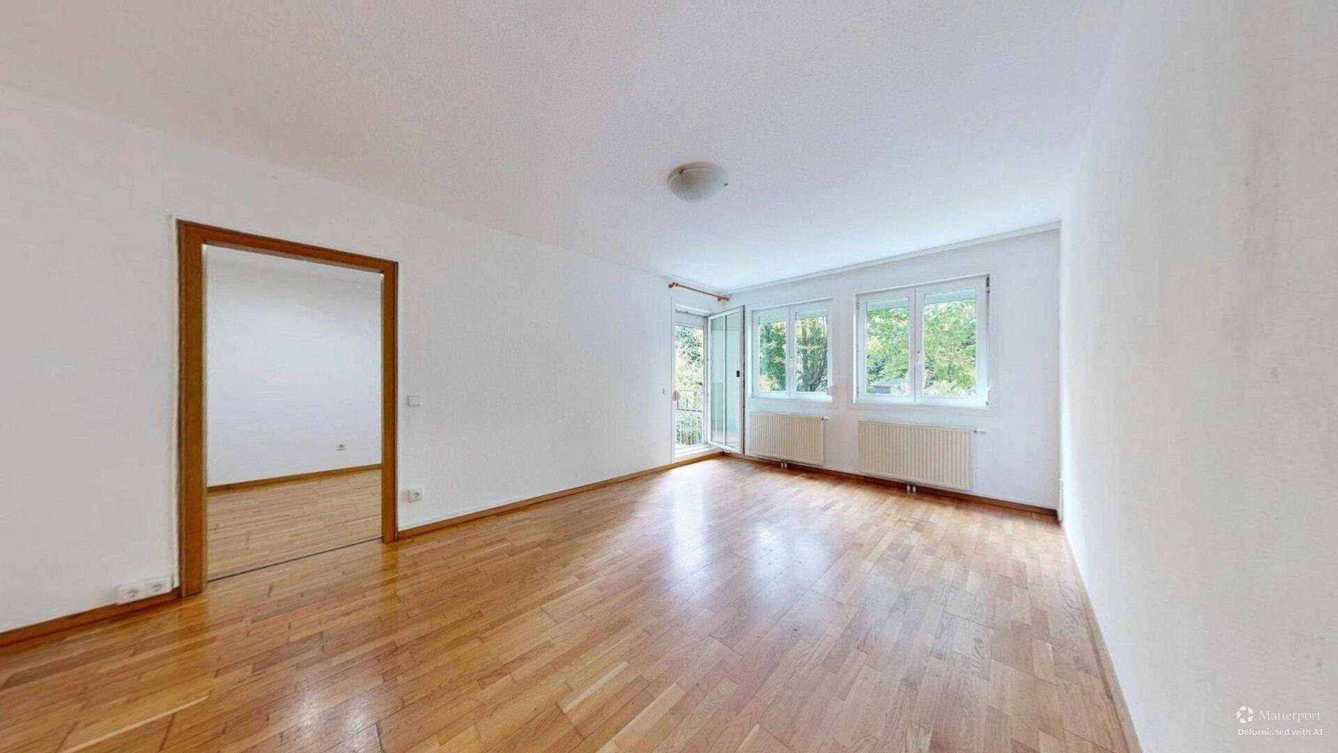 Helles, leeres Zimmer mit Parkettboden, weißen Wänden und Fenster mit Blick ins Grüne.