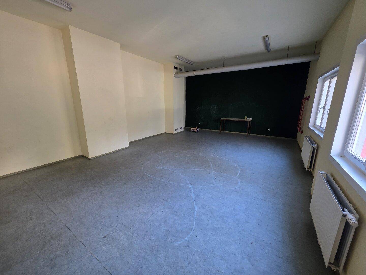 Großer, leerer Raum mit Betonboden, weißen Wänden und einer dunklen Akzentwand, ideal für verschiedene Nutzungen.