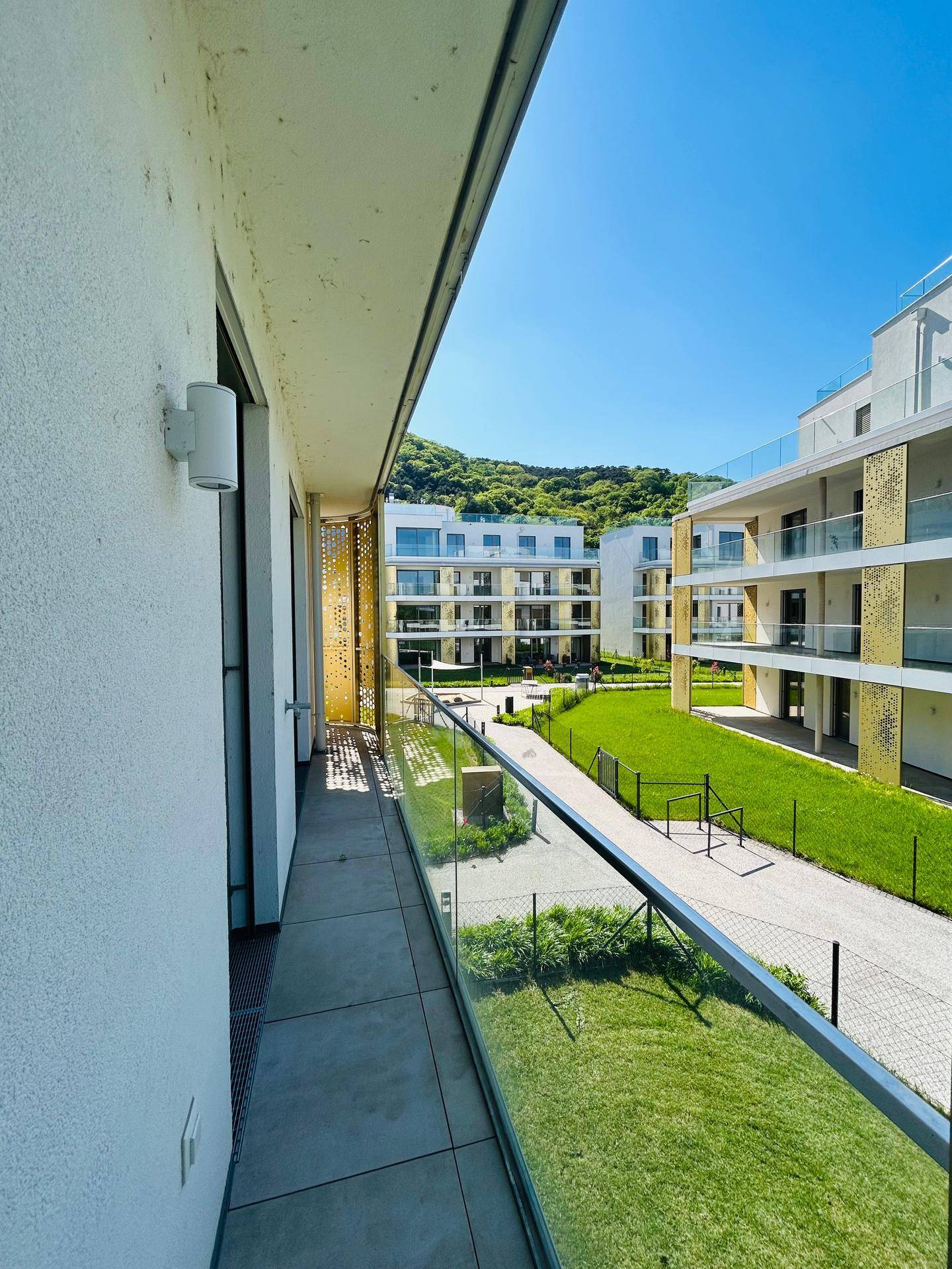 Sonniger Balkon mit Glasgeländer und Blick auf den grünen Innenhof und moderne Gebäude.