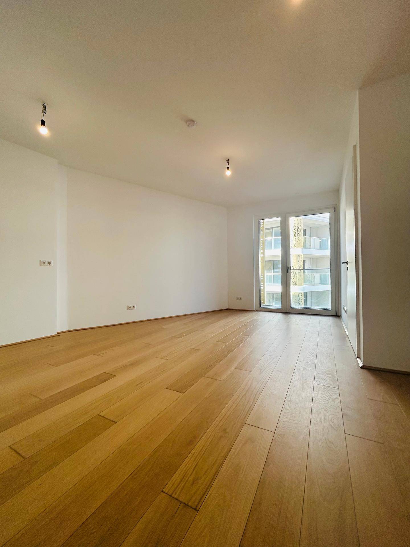 Lichtdurchfluteter Wohnraum mit hellem Holzboden und großen Fenstern, die zum Balkon führen.