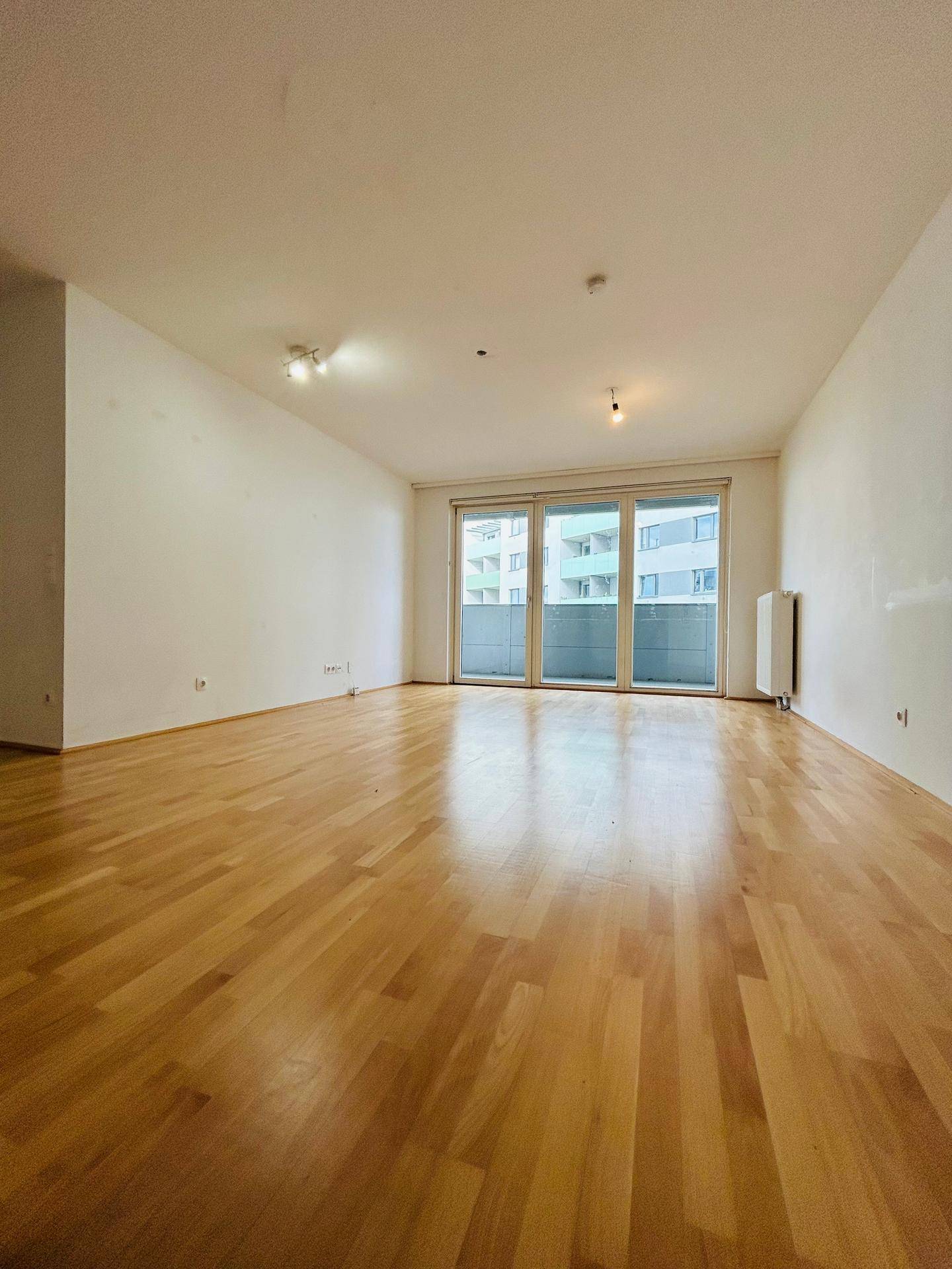 Helles, leeres Wohnzimmer mit glänzendem Parkettboden und Zugang zum Balkon.