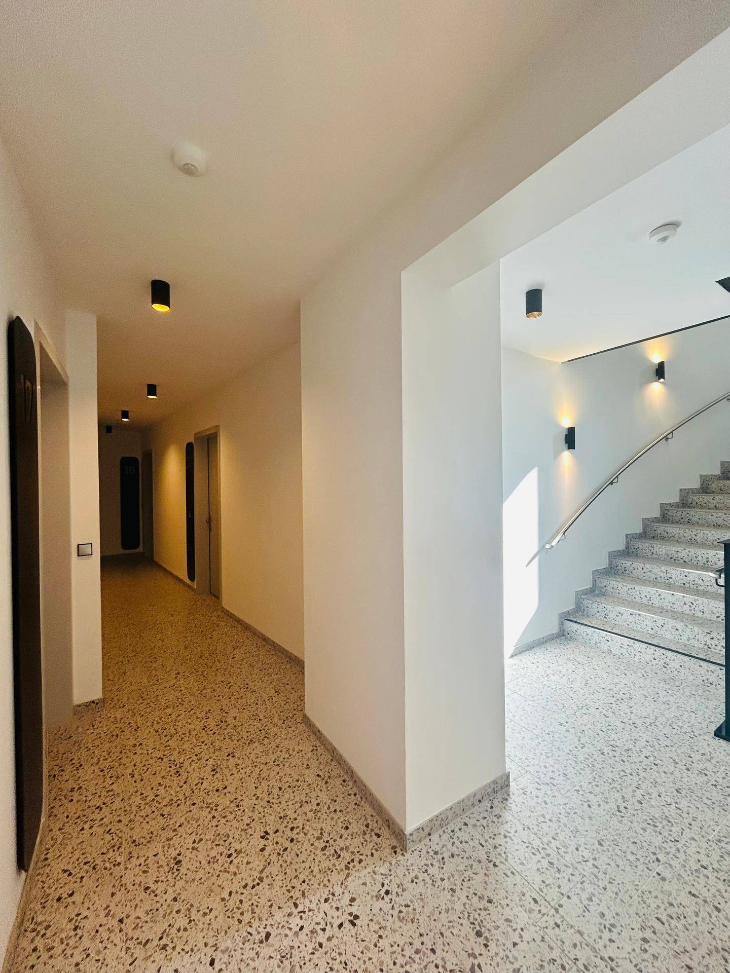 Heller Flur mit Treppenaufgang und moderner Beleuchtung, ausgestattet mit Terrazzoboden.