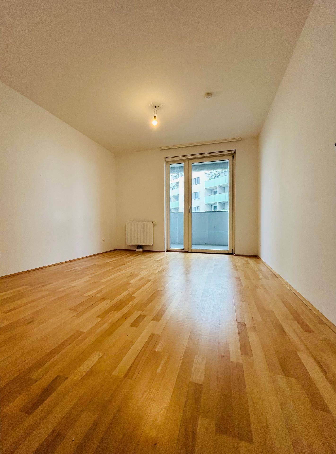 Leeres Zimmer mit hellem Parkettboden und großem Fenster, bereit zur Einrichtung.