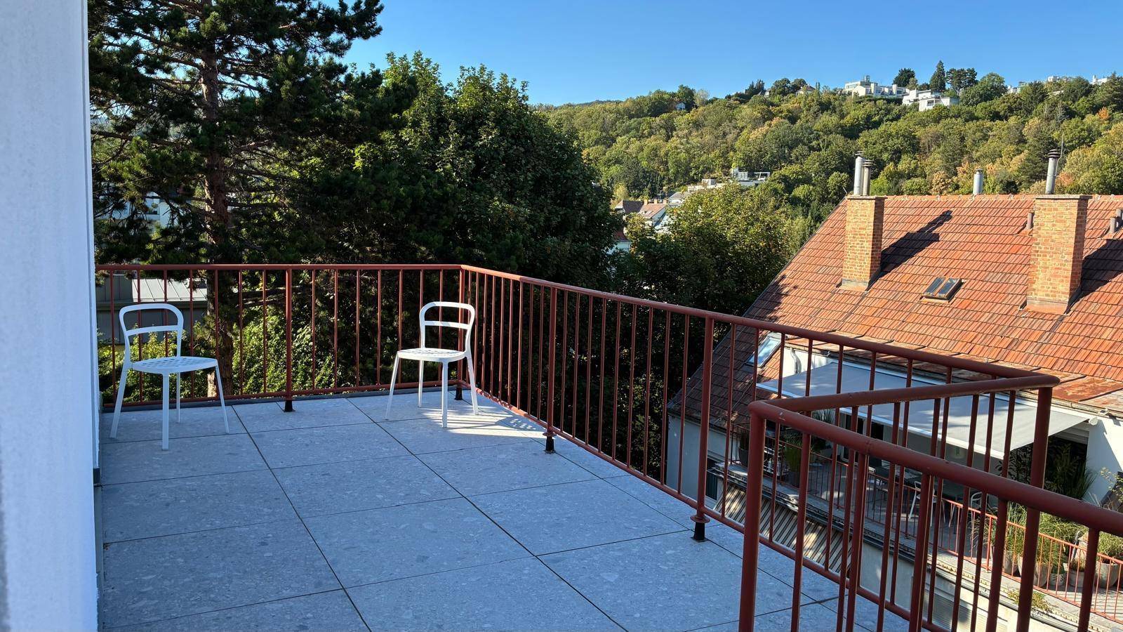 Sonniger Terrassenbereich mit zwei Stühlen, rotem Geländer und Blick auf die Umgebung.