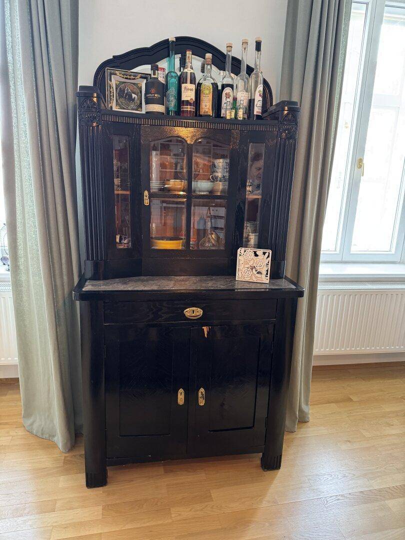 Antiker schwarzer Schrank mit Glasvitrine und Spirituosen zwischen zwei hohen Fenstern.