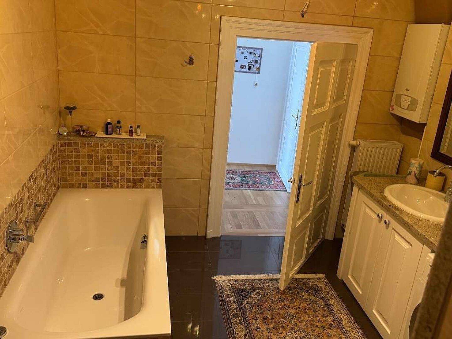 Badezimmer mit Badewanne, Fliesenboden und Zugang zu einem weiteren Raum.