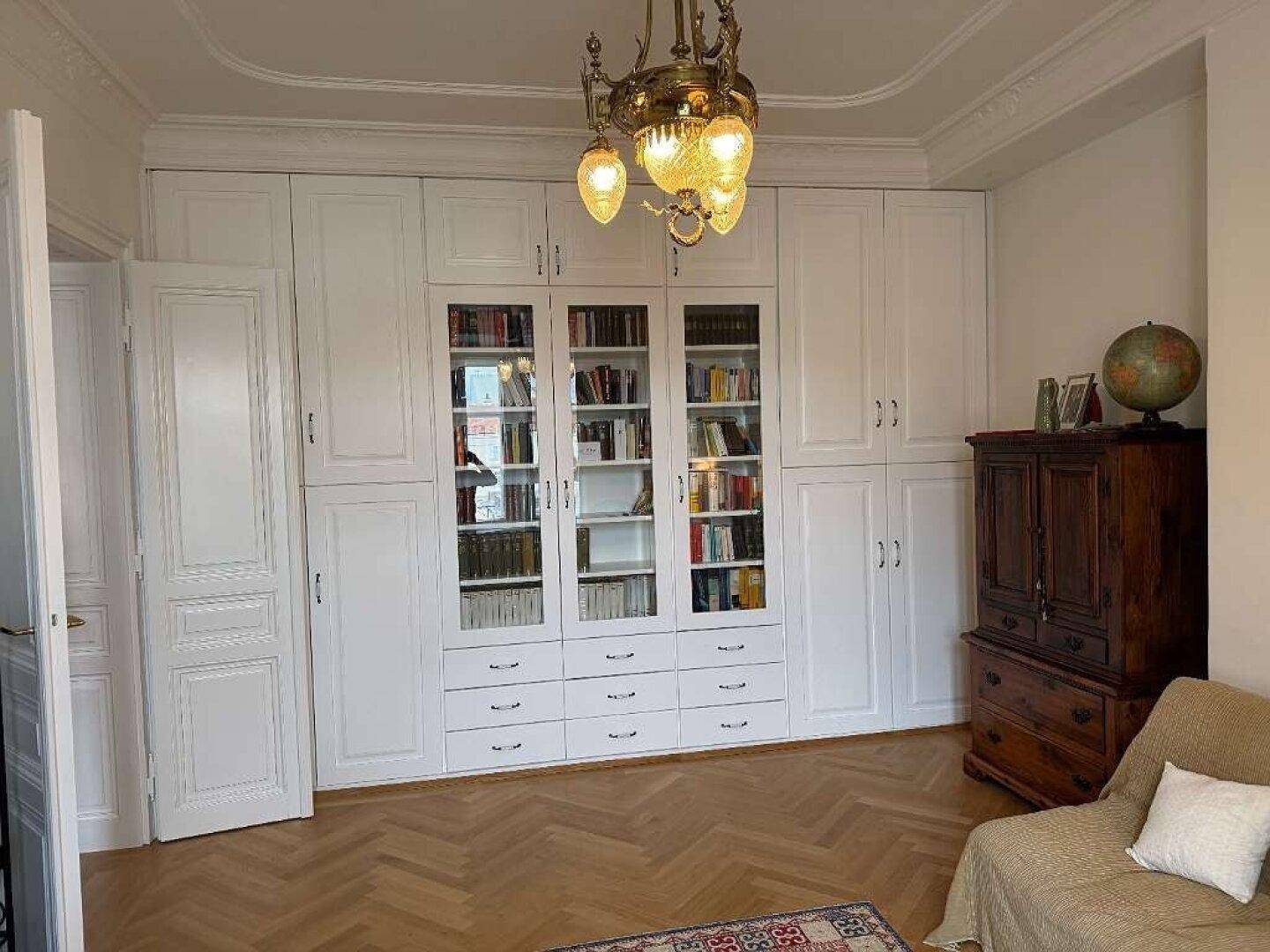 Wohnzimmer mit raumhohen weißen Schränken, Fischgrätparkett und einem Kronleuchter.