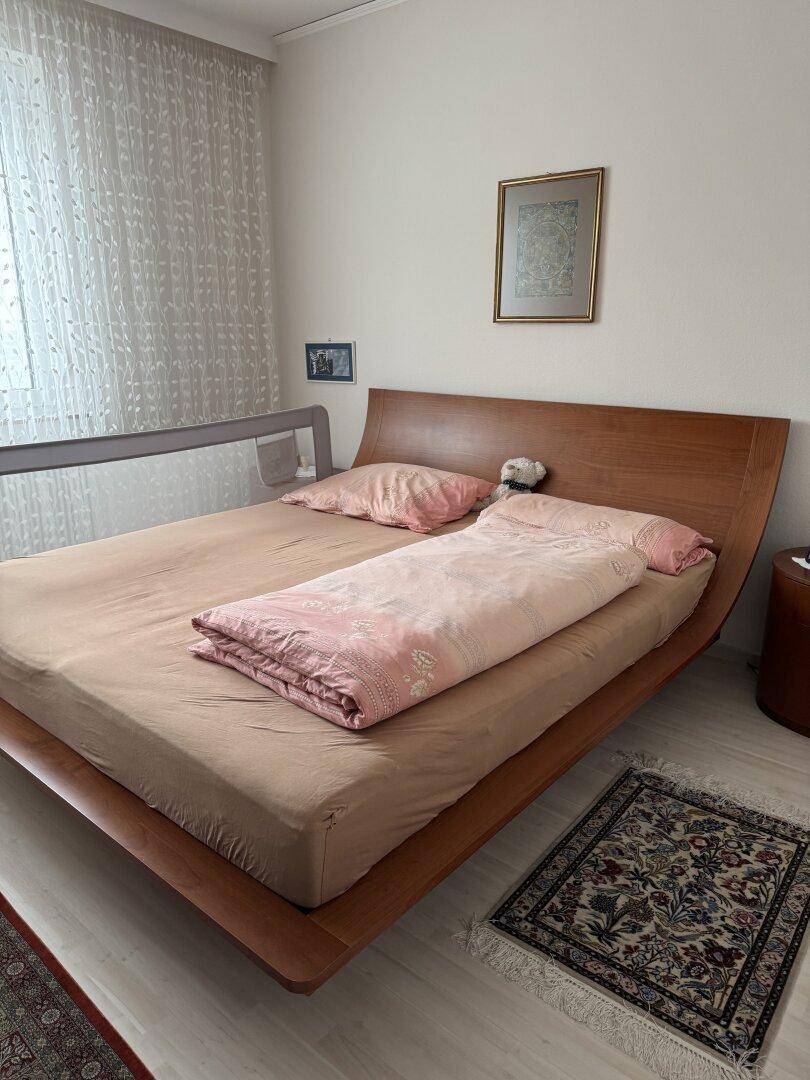 Schlafzimmer mit modernem Holzbett, Teppich und hellem Fensterbereich.