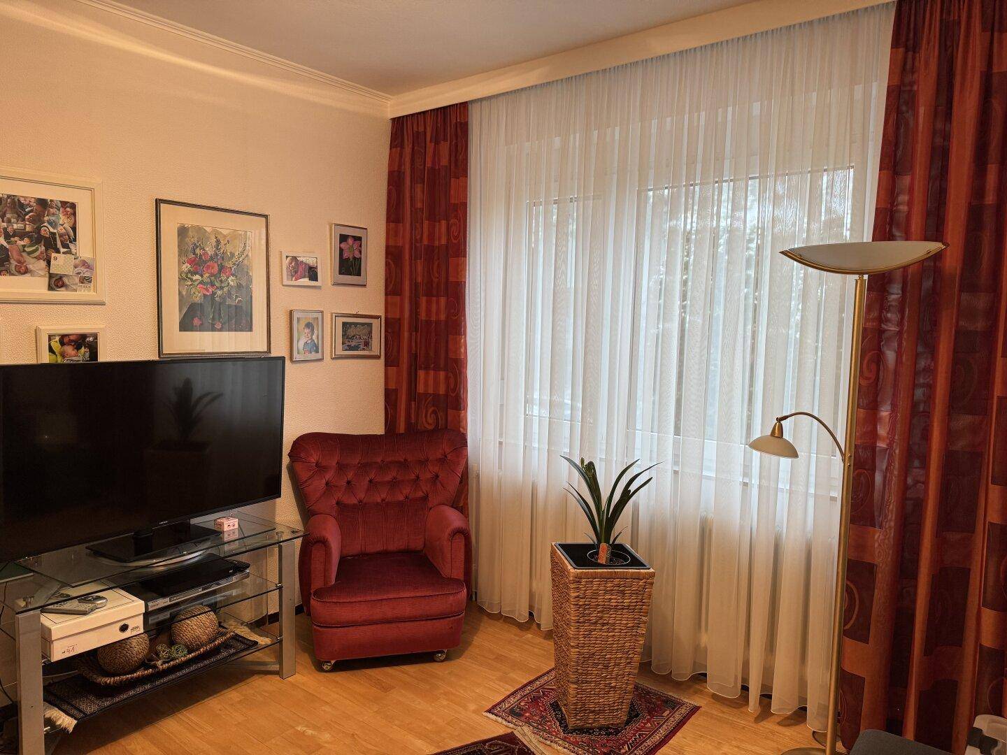 Gemütliches Wohnzimmer mit Fernseher, rotem Sessel und großen Fenstern mit Vorhängen.
