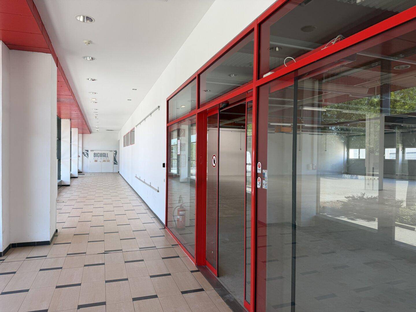 Langer Innenflur mit Fliesenboden und leeren Geschäftsräumen mit roten Glasfronten.