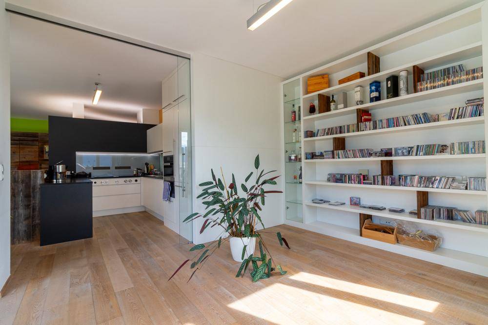 Heller Raum mit Holzboden und Blick auf die moderne Küche durch eine Glastür.