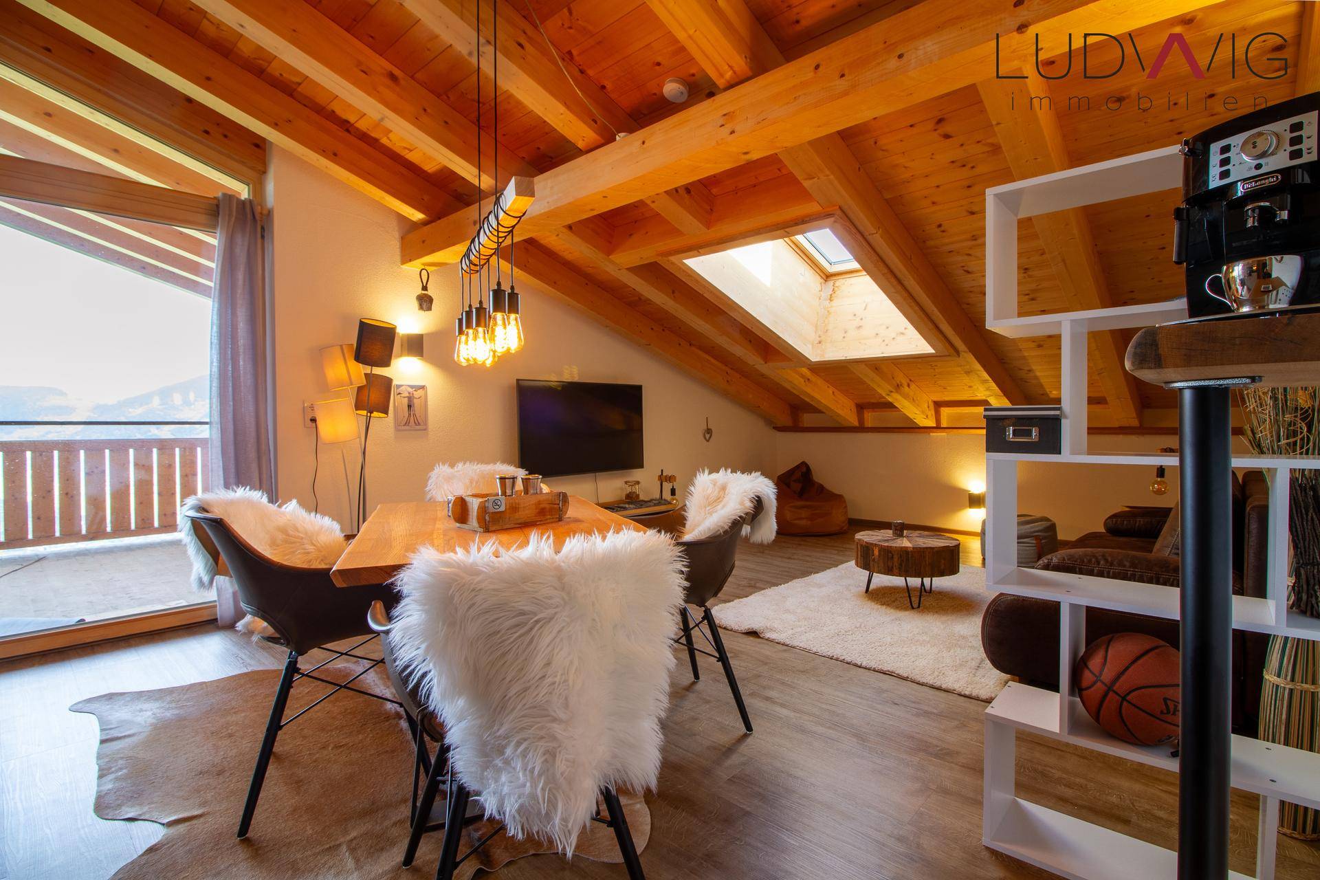 Gemütlicher Wohn-Essbereich im Dachgeschoss mit Holzbalken, Dachfenster und Zugang zum Balkon mit Bergblick.