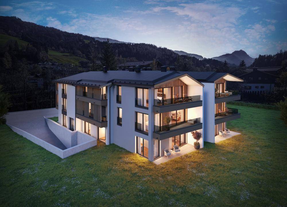 Modernes Mehrfamilienhaus mit beleuchteten Innenräumen und Balkonen vor einer Bergkulisse bei Nacht.
