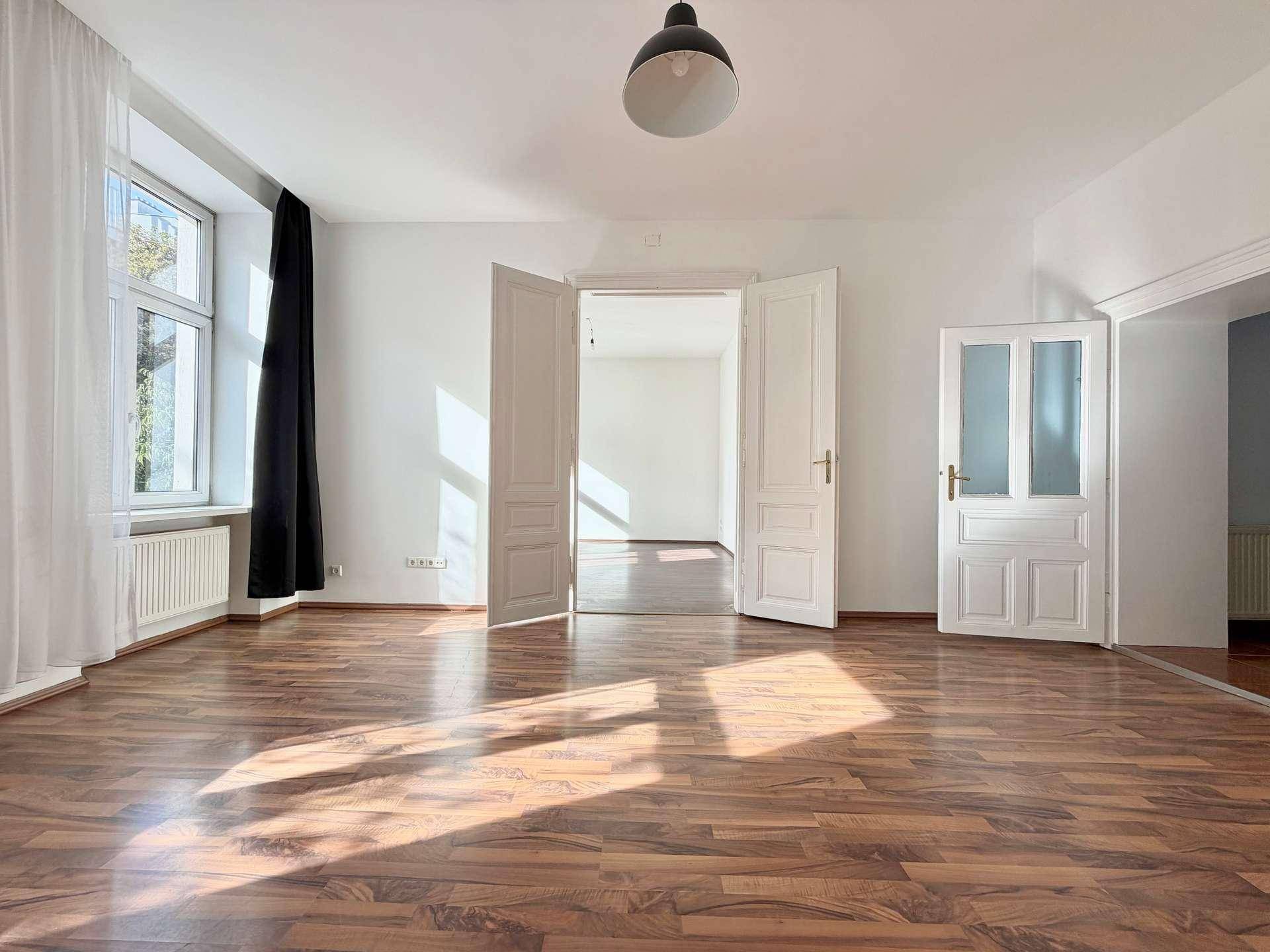 Geräumiges Wohnzimmer mit Laminatboden, hohen Decken und doppelten Flügeltüren zu einem weiteren Raum.