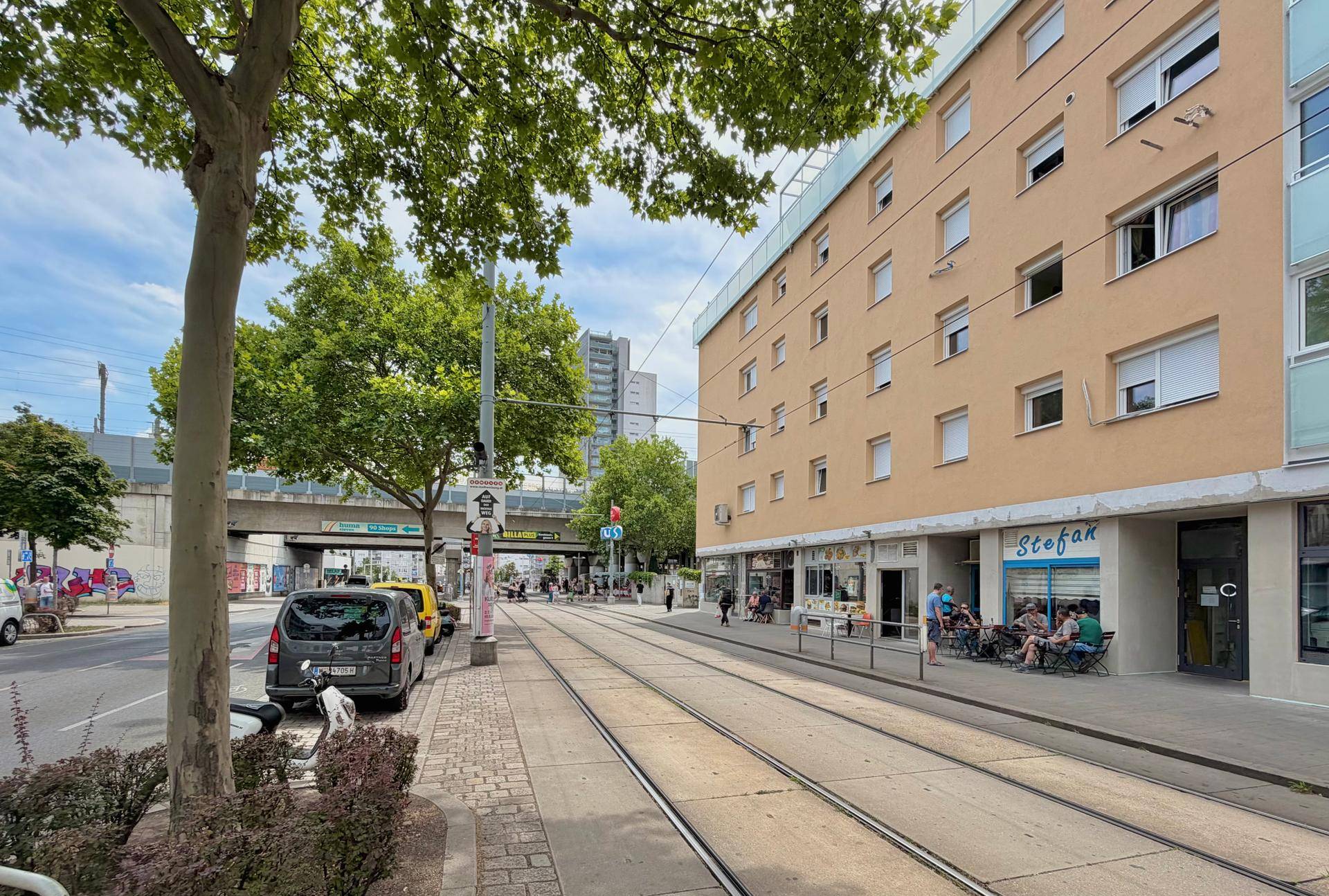 Belebtes Stadtbild mit Straßenbahnschienen, Geschäften und einem Wohngebäude unter blauem Himmel.