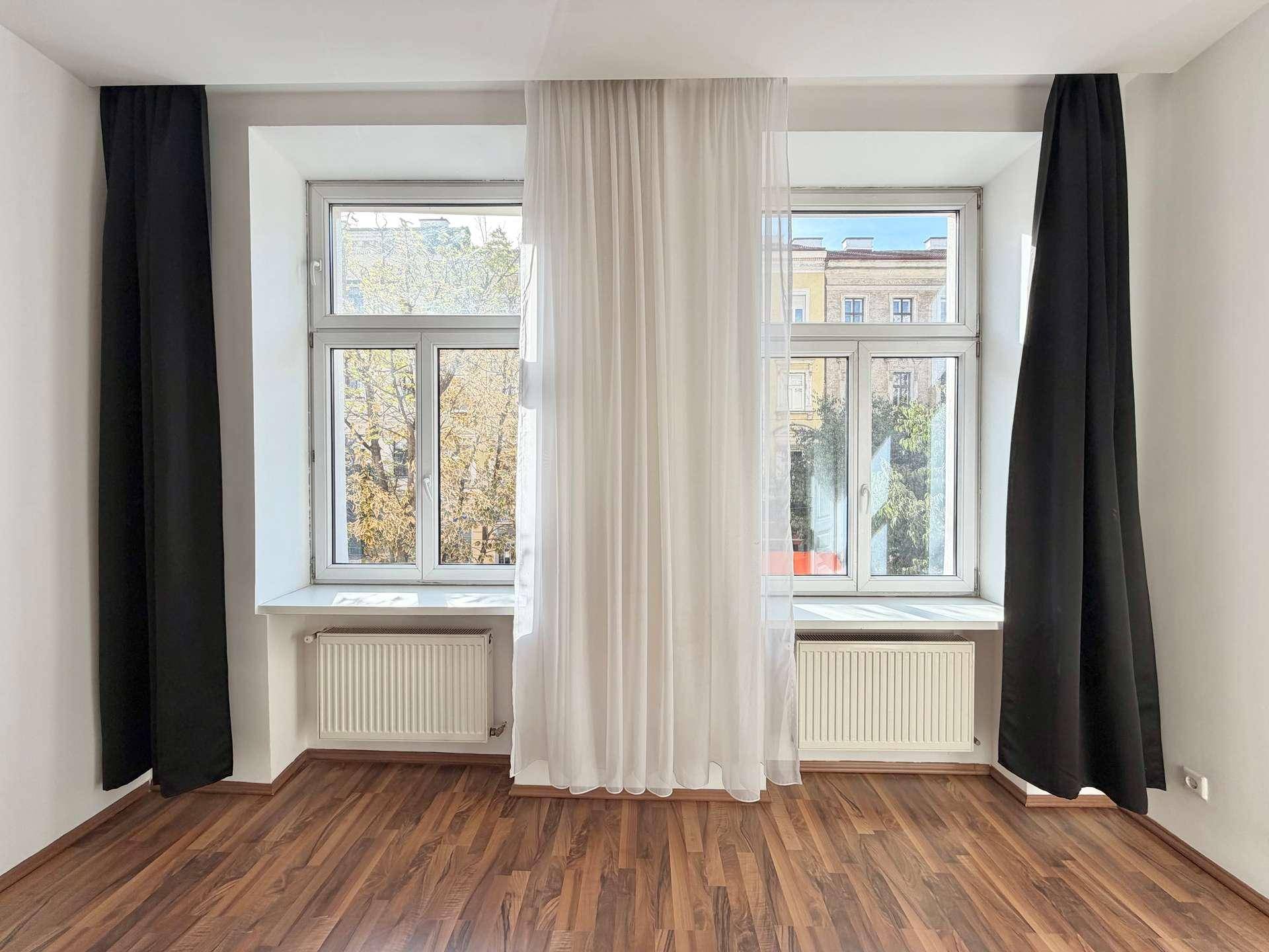 Nahaufnahme der Fenster im Wohnzimmer mit weißen Vorhängen und Blick auf Bäume und Gebäude.