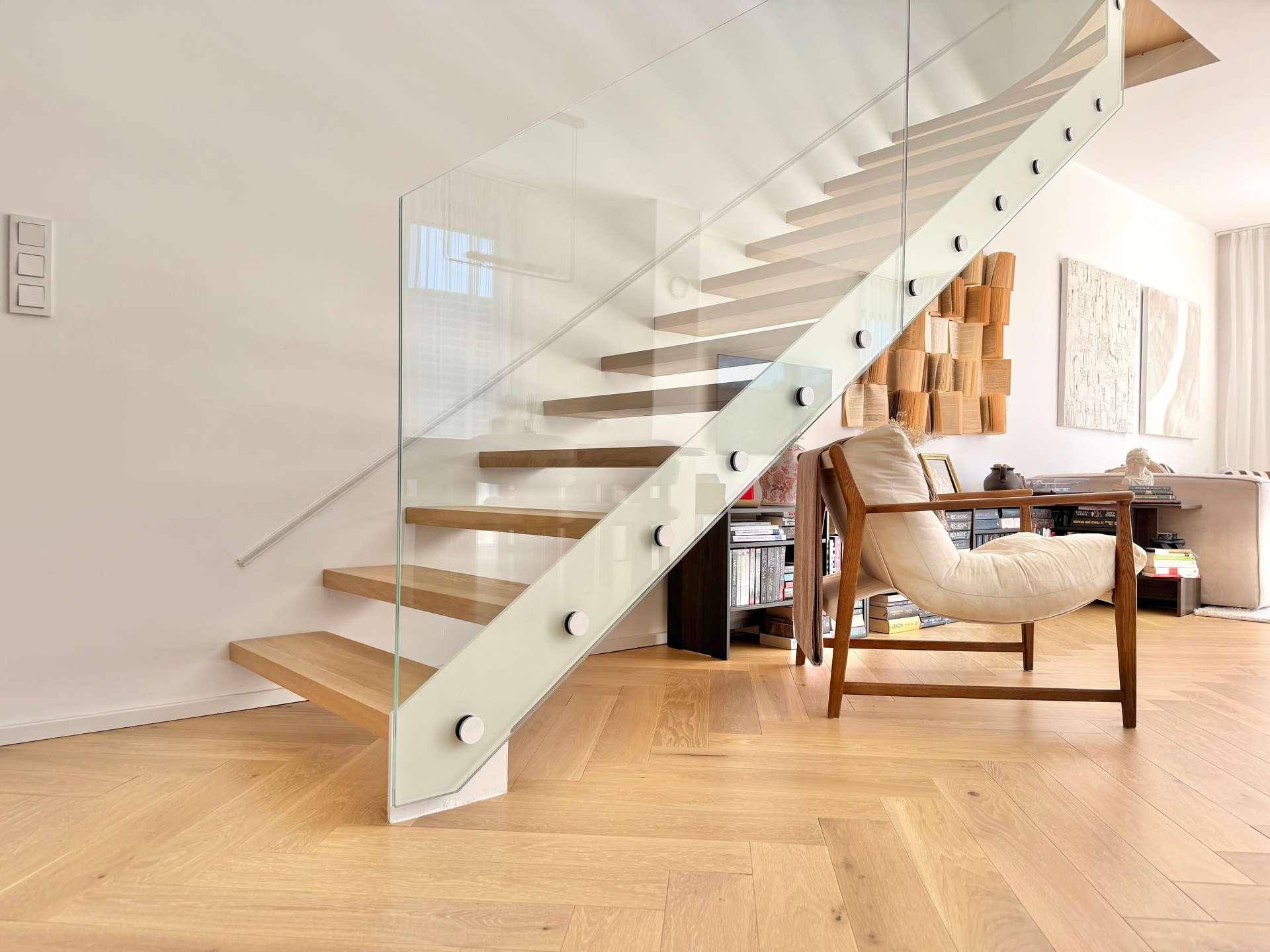 Moderne Treppe mit Glasgeländer und Holzstufen, dazu Fischgrätparkett im Wohnbereich.