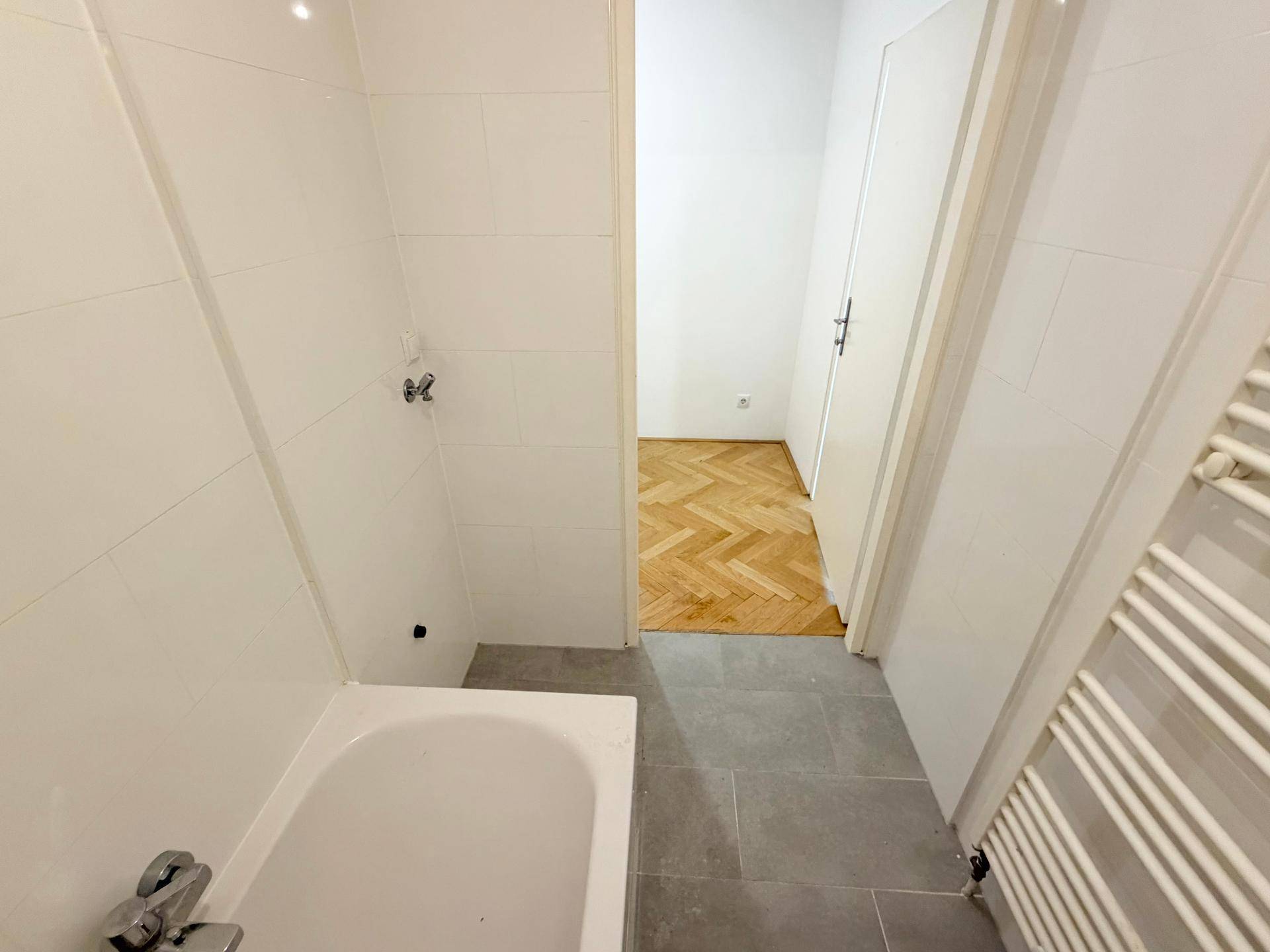 Badezimmer mit Badewanne und Fliesen, das einen Übergang zu einem Zimmer mit Parkettboden bietet.