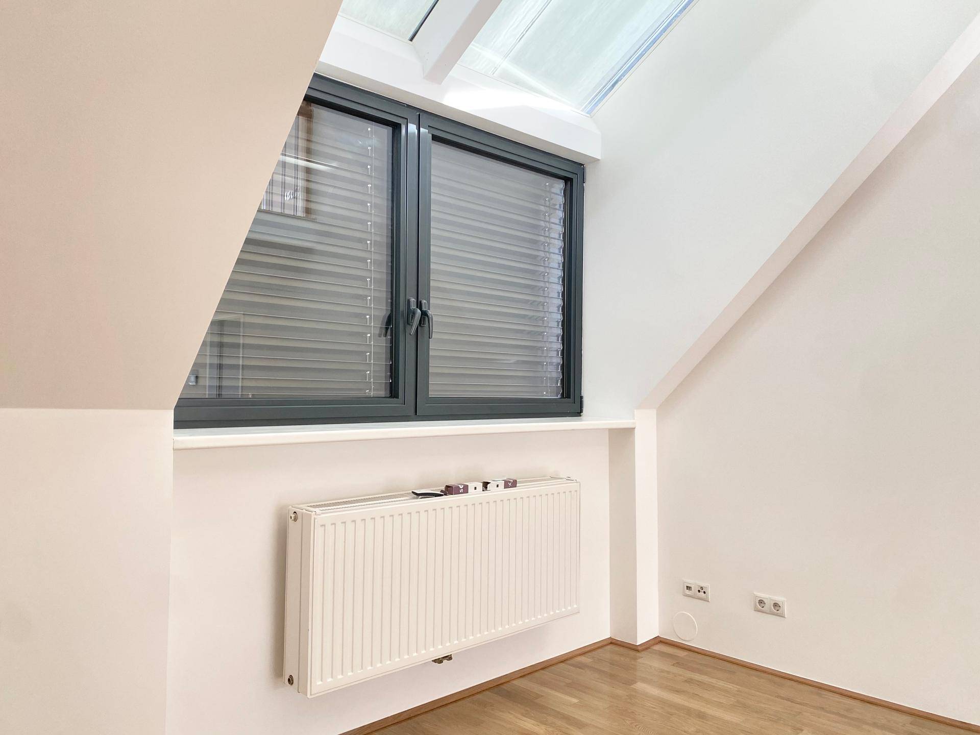 Heller Raum mit schrägen Wänden, Parkettboden und einem Fenster mit Jalousien.