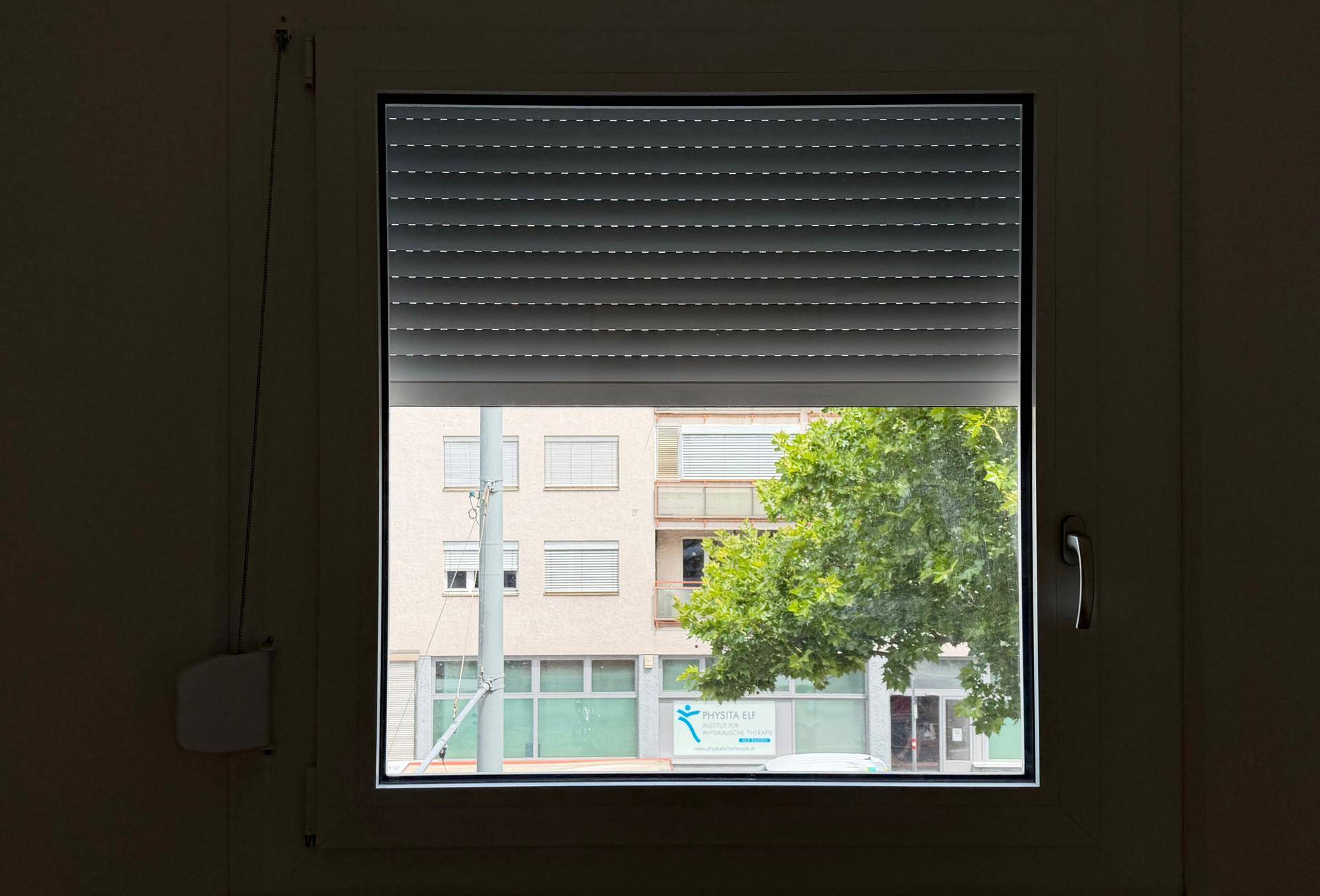 Fenster mit teilweise heruntergelassenem Rollladen, das den Blick auf die Straße freigibt.