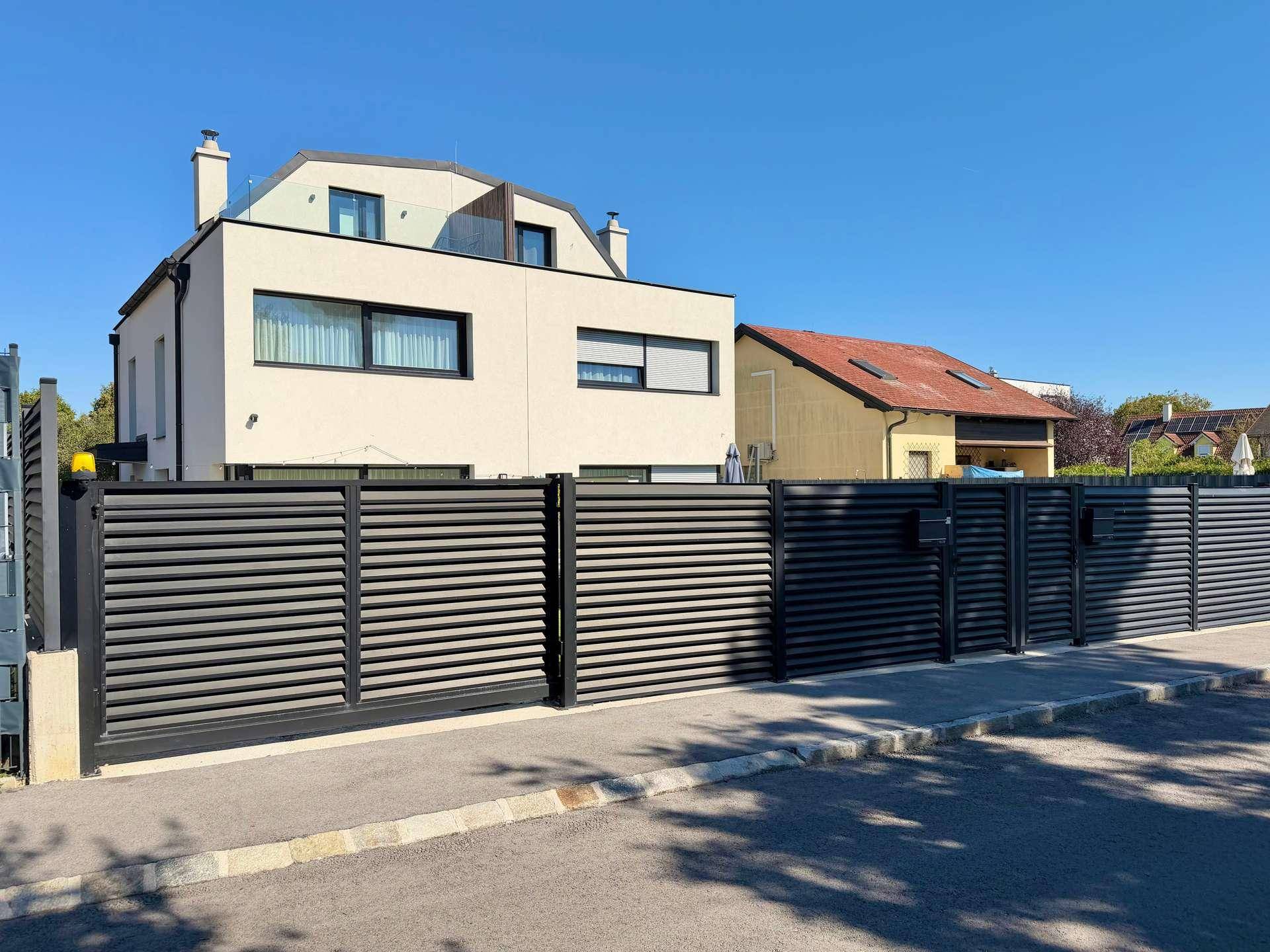 Moderne Hausfassade mit dunklem Zaun und klaren Linien unter blauem Himmel.