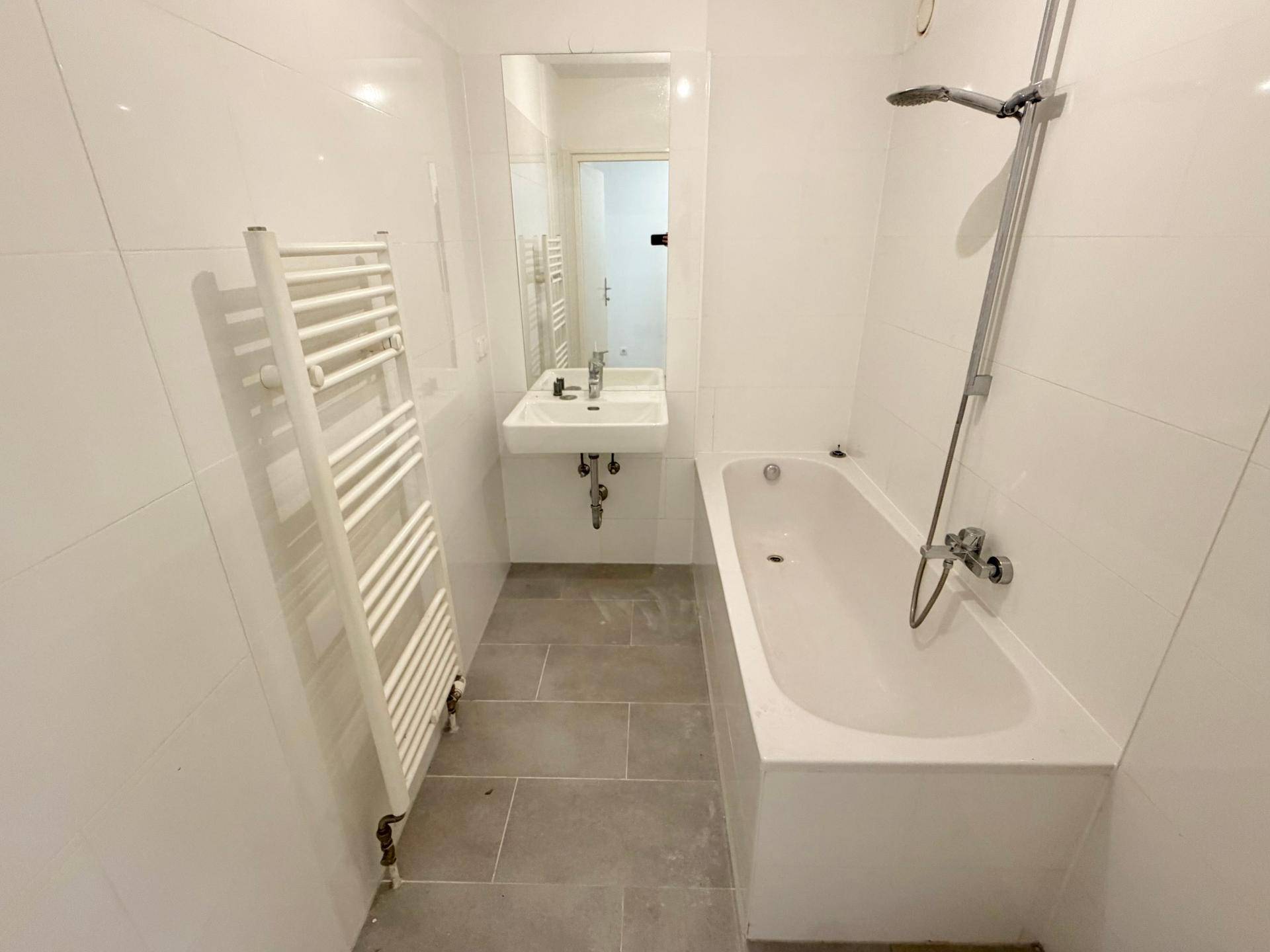 Modernes Badezimmer mit weißen Wandfliesen, grauen Bodenfliesen, Badewanne und Handtuchheizkörper.