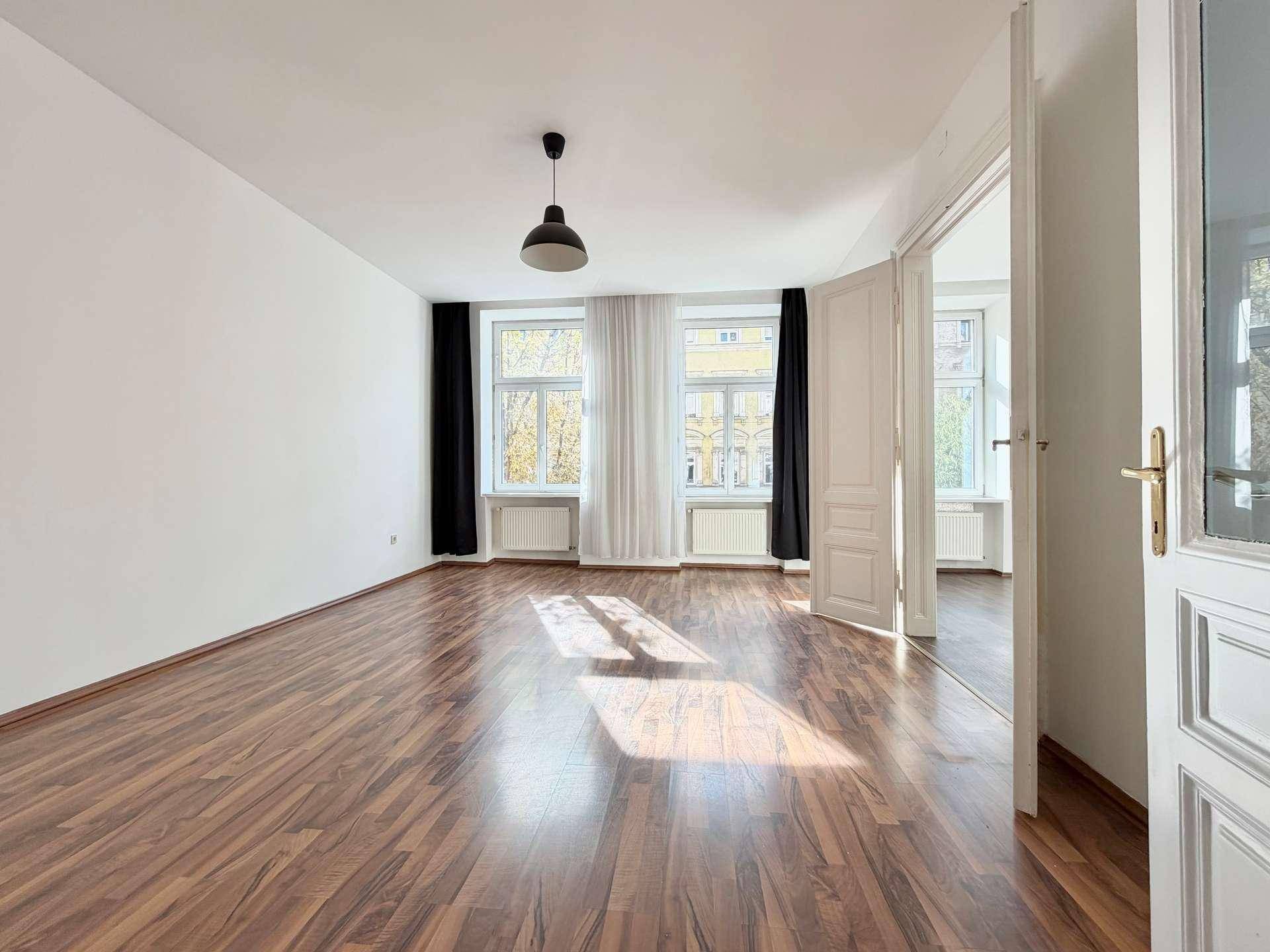 Großes Wohnzimmer mit Laminatboden, hohen Decken und viel Tageslicht durch mehrere Fenster.