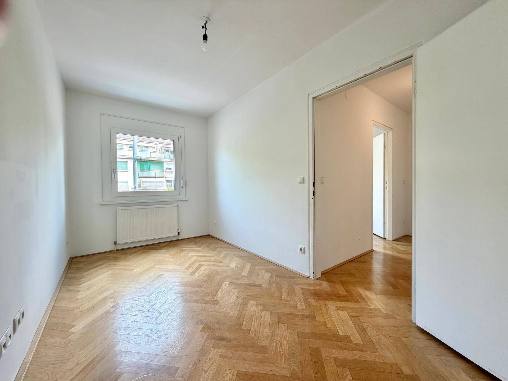 Helles Zimmer mit Fischgrätparkett und einem Fenster, das den Blick nach draußen freigibt.