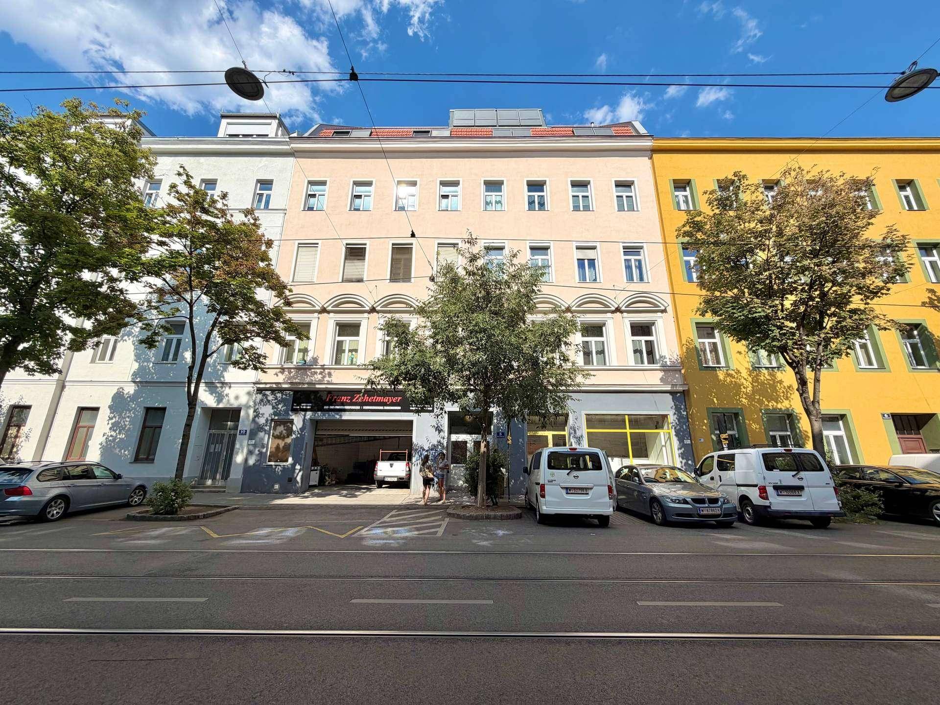 Fassade eines Wohngebäudes an einer belebten Straße mit geparkten Fahrzeugen und Geschäften im Erdgeschoss.